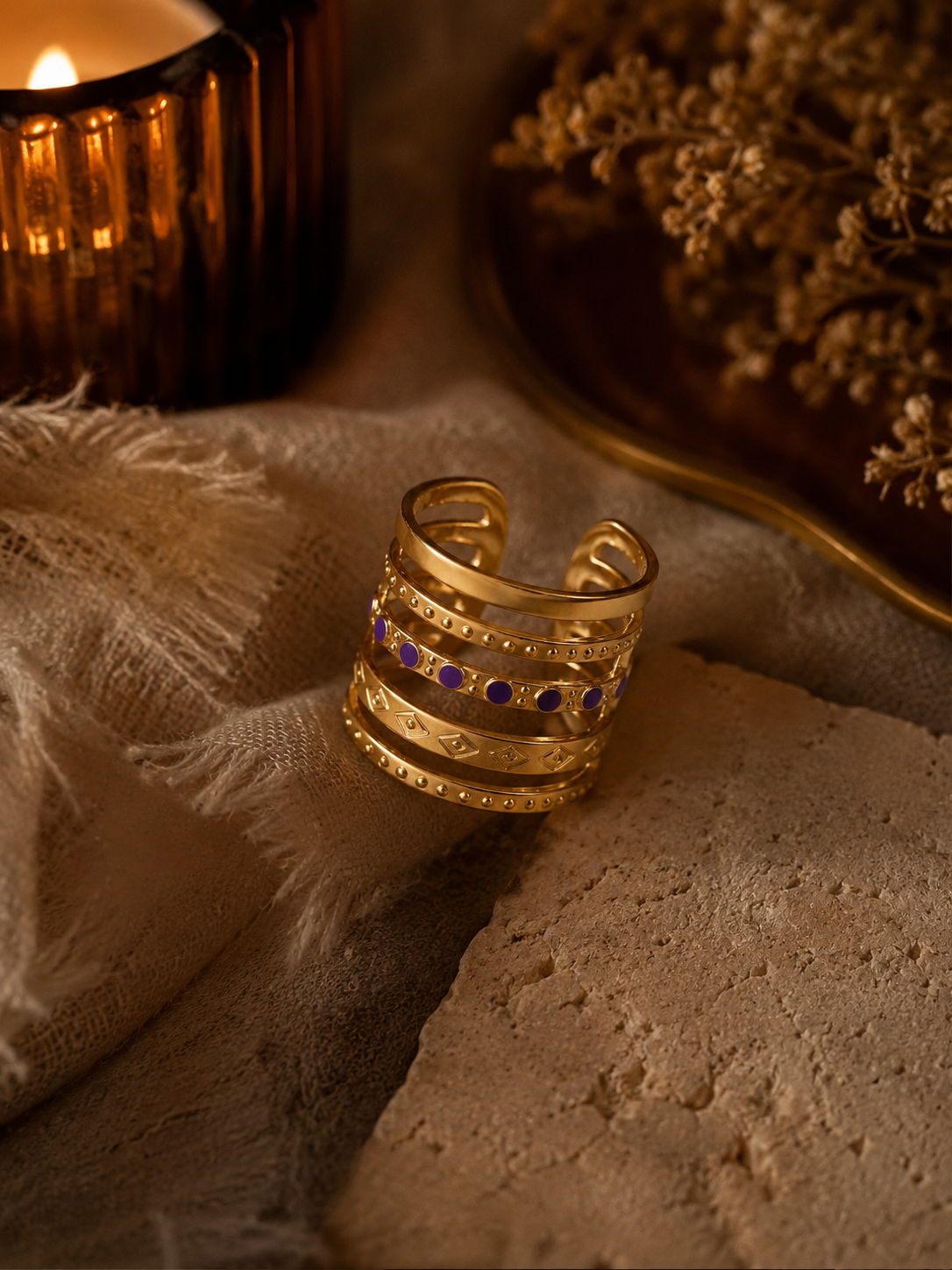 Bague Valéria dorée avec détails violets posée sur pierre beige et tissu naturel dans une ambiance cosy et chaleureuse – Bella Cosa