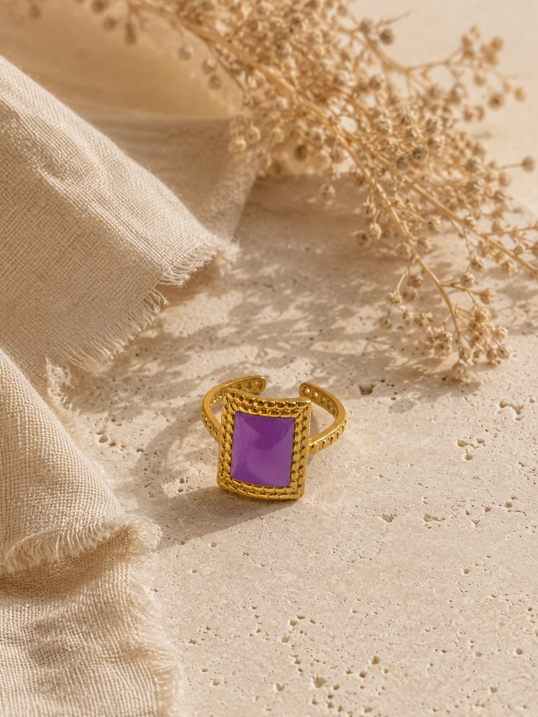Bague Vélina dorée et violette posée sur travertin beige et tissu naturel dans une ambiance chaleureuse et raffinée – Bella Cosa