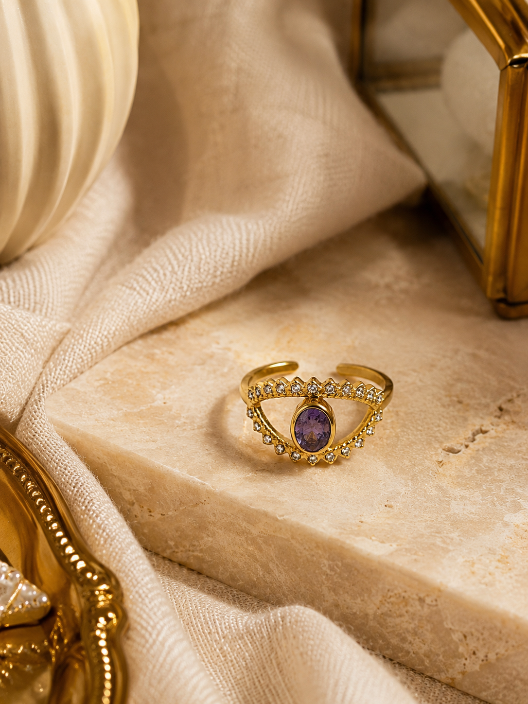 Bague Victoria dorée avec pierre violette posée sur travertin beige et tissu naturel dans une ambiance chic et chaleureuse – Bella Cosa