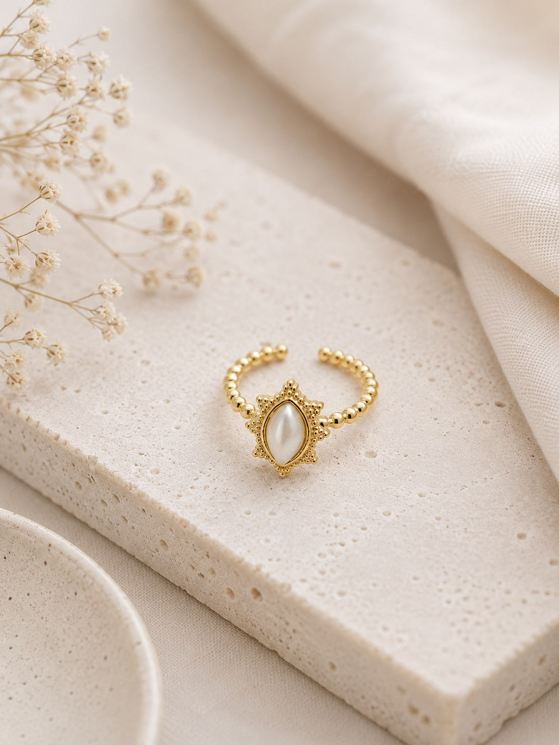Bague Alba dorée avec pierre nacrée posée sur travertin beige et lin naturel dans une ambiance douce et chaleureuse – Bella Cosa
