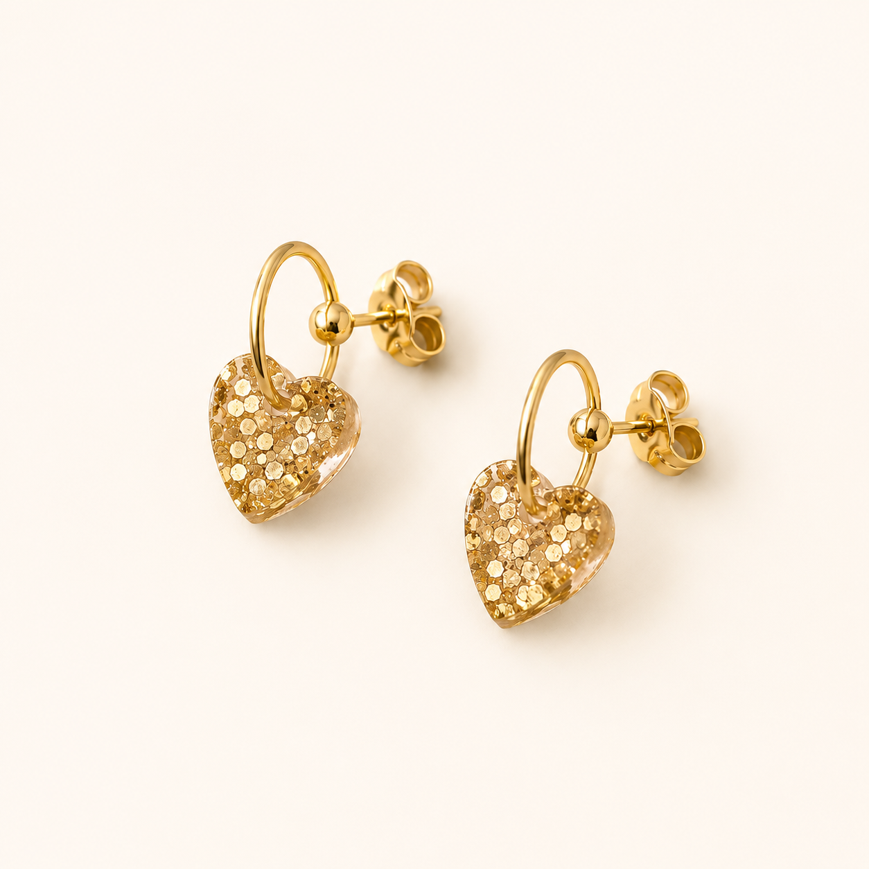 Boucles d’oreilles Cassie dorées avec cœur en résine pailletée sur fond beige clair élégant