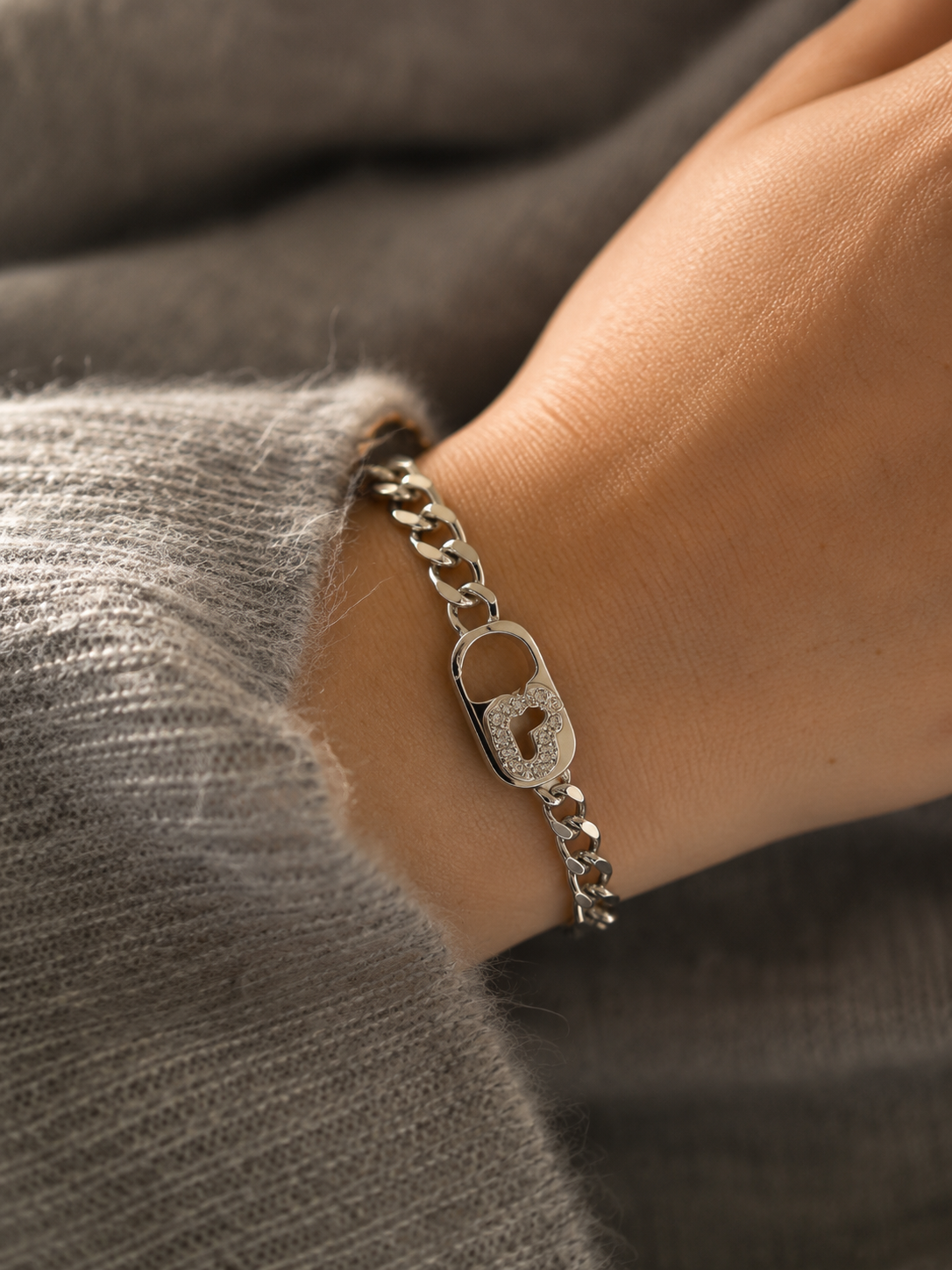 Bracelet Gizem argenté à grosses mailles avec fermoir cœur serti de zirconiums porté sur un poignet féminin en maille grise douce