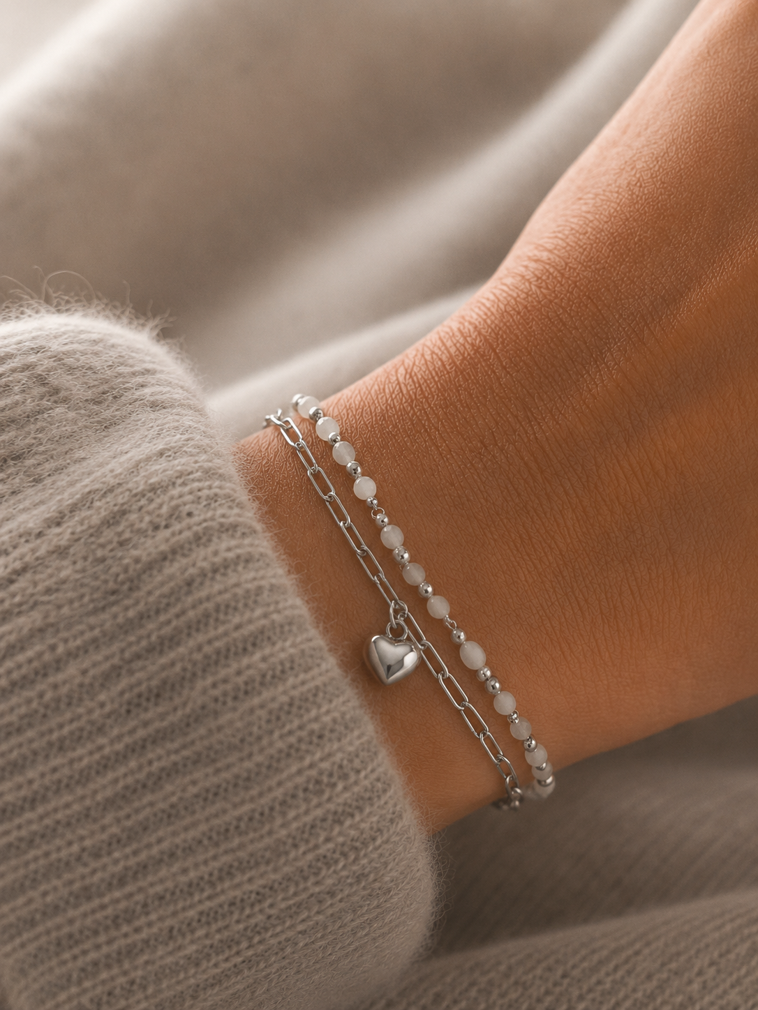 Bracelet April argenté double rang avec perles blanches et pendentif cœur porté sur un poignet féminin en maille douce beige taupe