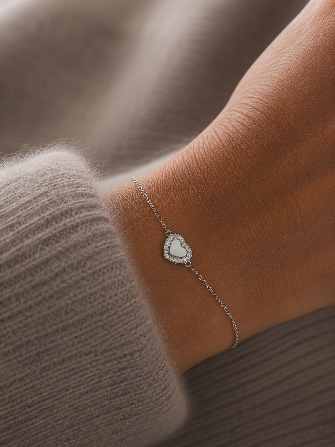 Bracelet Noa argenté avec cœur blanc nacré et zirconiums porté sur un poignet féminin en maille douce beige taupe