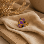 Bague Vera dorée et violette posée sur pierre beige et tissu naturel dans une ambiance chaleureuse et raffinée – Bella Cosa
