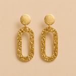 Boucles d’oreilles Clémentine dorées en résine pailletée sur fond beige élégant