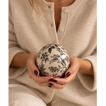 Boule décorative en grès écru à motifs floraux noirs tenue dans les mains.