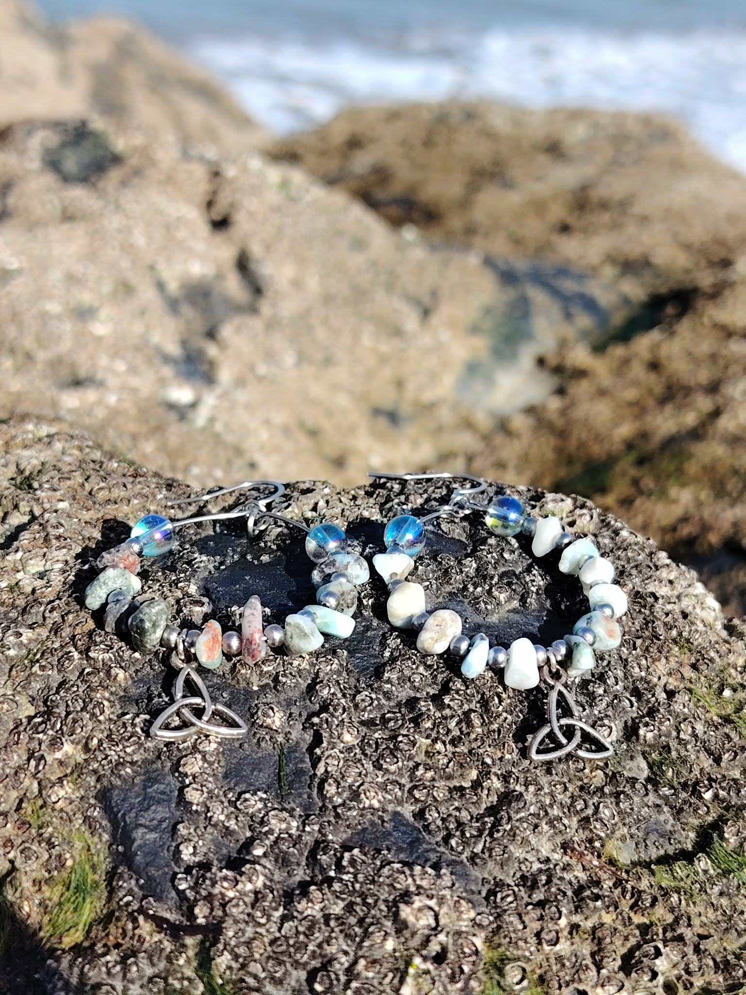 Boucles d’oreilles Magic Gaïa en larimar et aqua aura posées sur un rocher granitique en Bretagne, face à la mer floue, avec perles bleu ciel et blanches, triquetra argentée, évoquant douceur, intuition et apaisement.