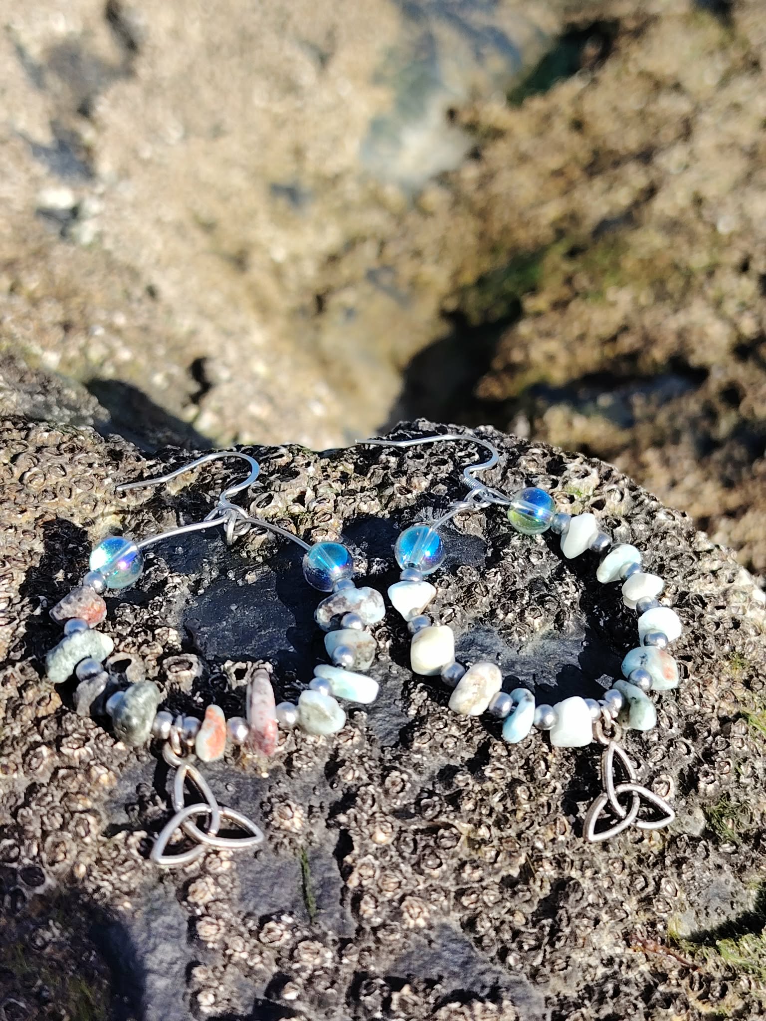 Boucles d’oreilles Magic Gaïa en larimar et aqua aura posées sur rocher granitique en Bretagne, perles bleu ciel et blanches en cercle avec triquetra, ambiance minérale naturelle évoquant douceur, intuition et apaisement