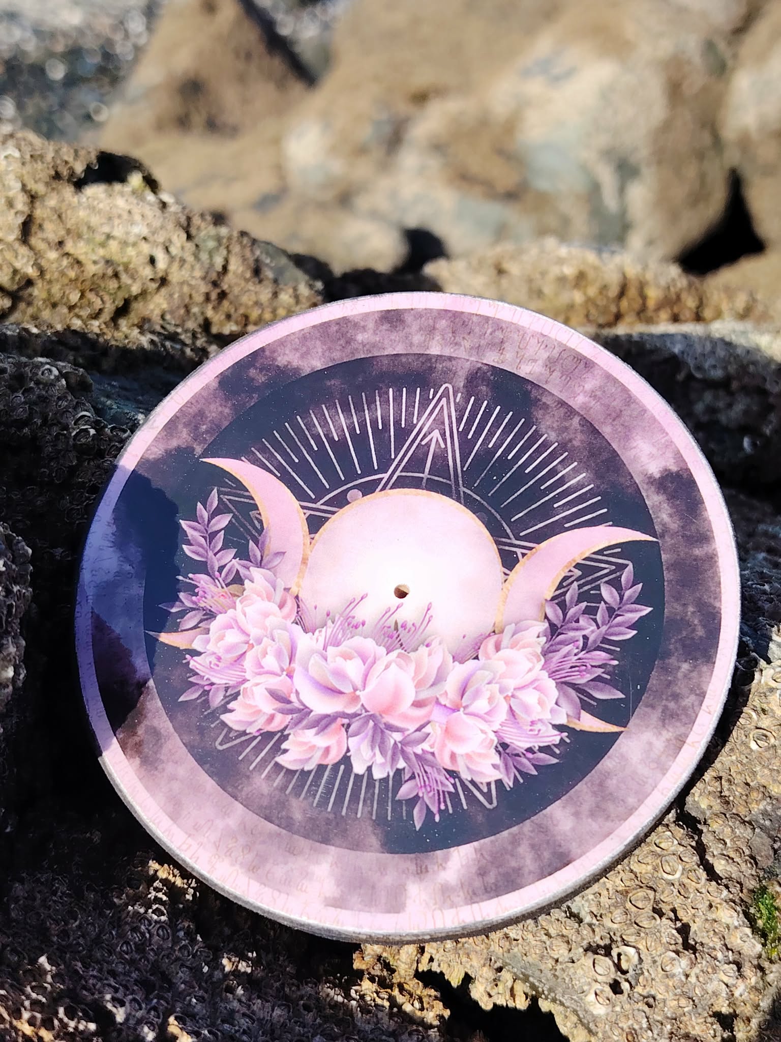 Brûleur d’encens rond violet motif Triple Lune posé sur rocher près de la mer en Bretagne; fumigation, atmosphère apaisante et protection symbolique.