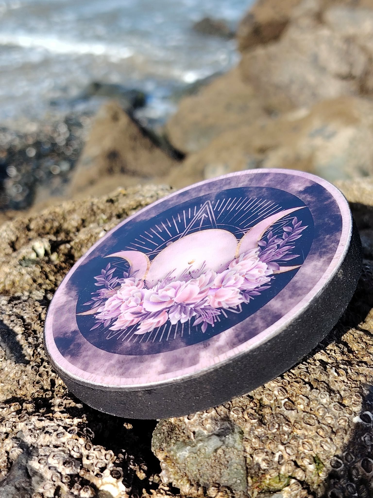 Brûleur d’encens rond violet Triple Lune sur rocher face à la mer en Bretagne; fumigation, ambiance apaisante et protection symbolique.