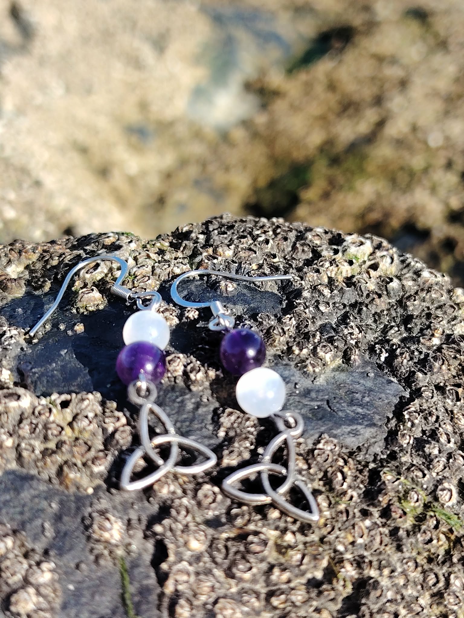 Boucles d’oreilles elfiques triquetra en améthyste et sélénite, posées sur rocher côtier face à la mer en Bretagne, pour apaisement et clarté