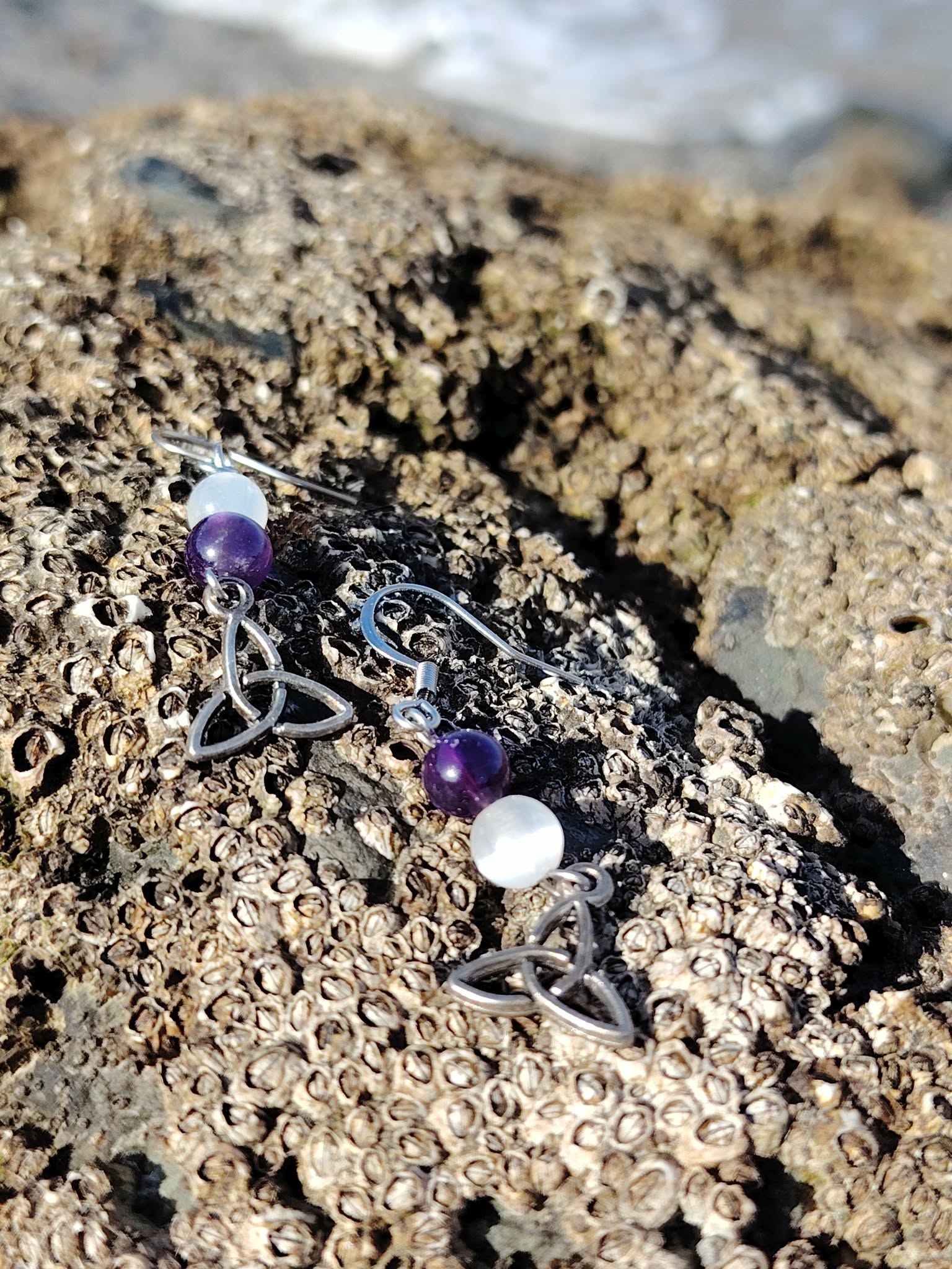 Boucles d’oreilles elfiques triquetra améthyste et sélénite posées sur rocher au bord de mer en Bretagne, création Magic Gaïa, pour apaiser et éclaircir l’esprit.