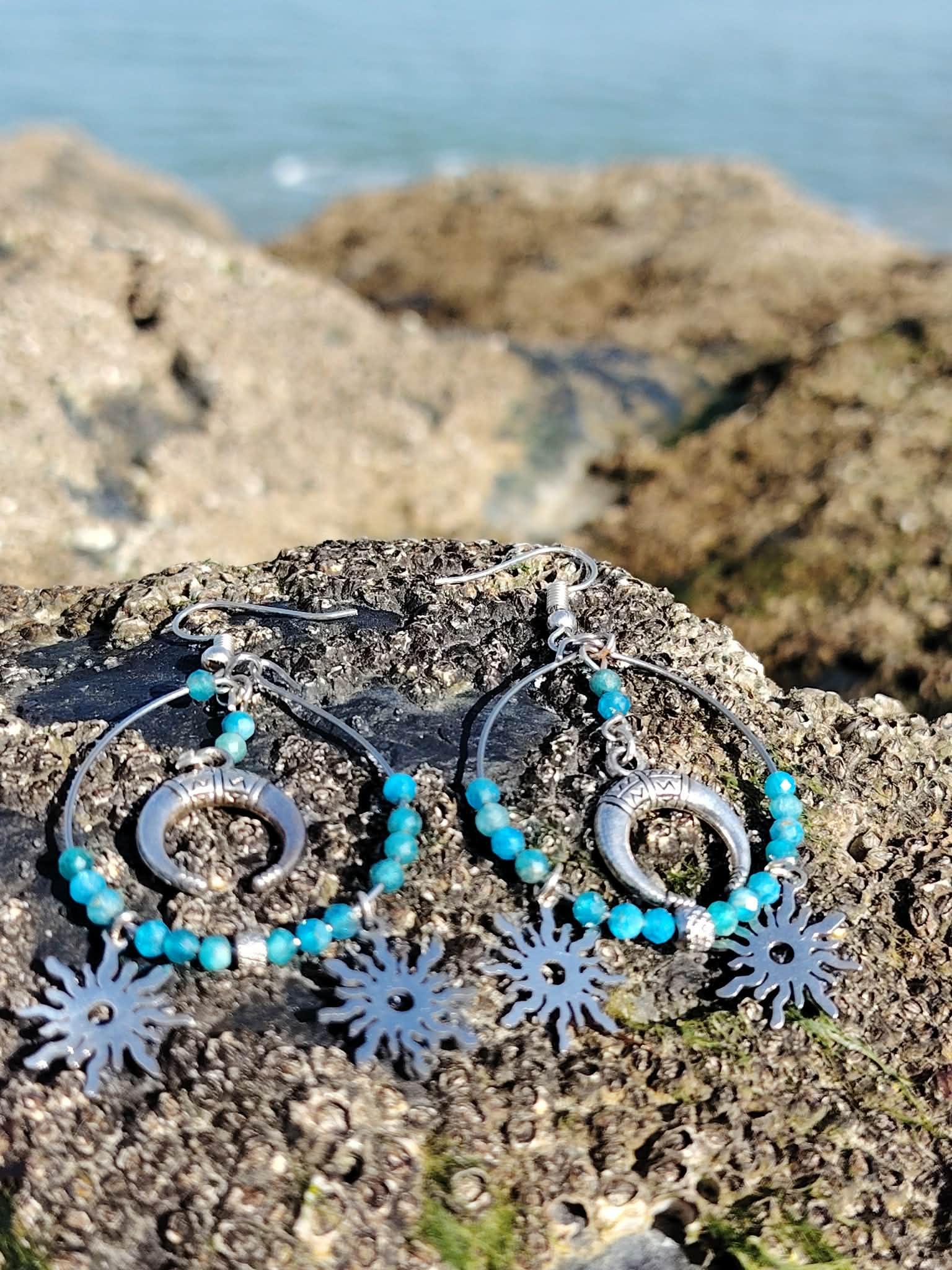 Boucles d’oreilles Atlante en apatite, création Magic Gaïa, sur rocher breton avec mer en arrière-plan, pour calme intérieur et communication posée.