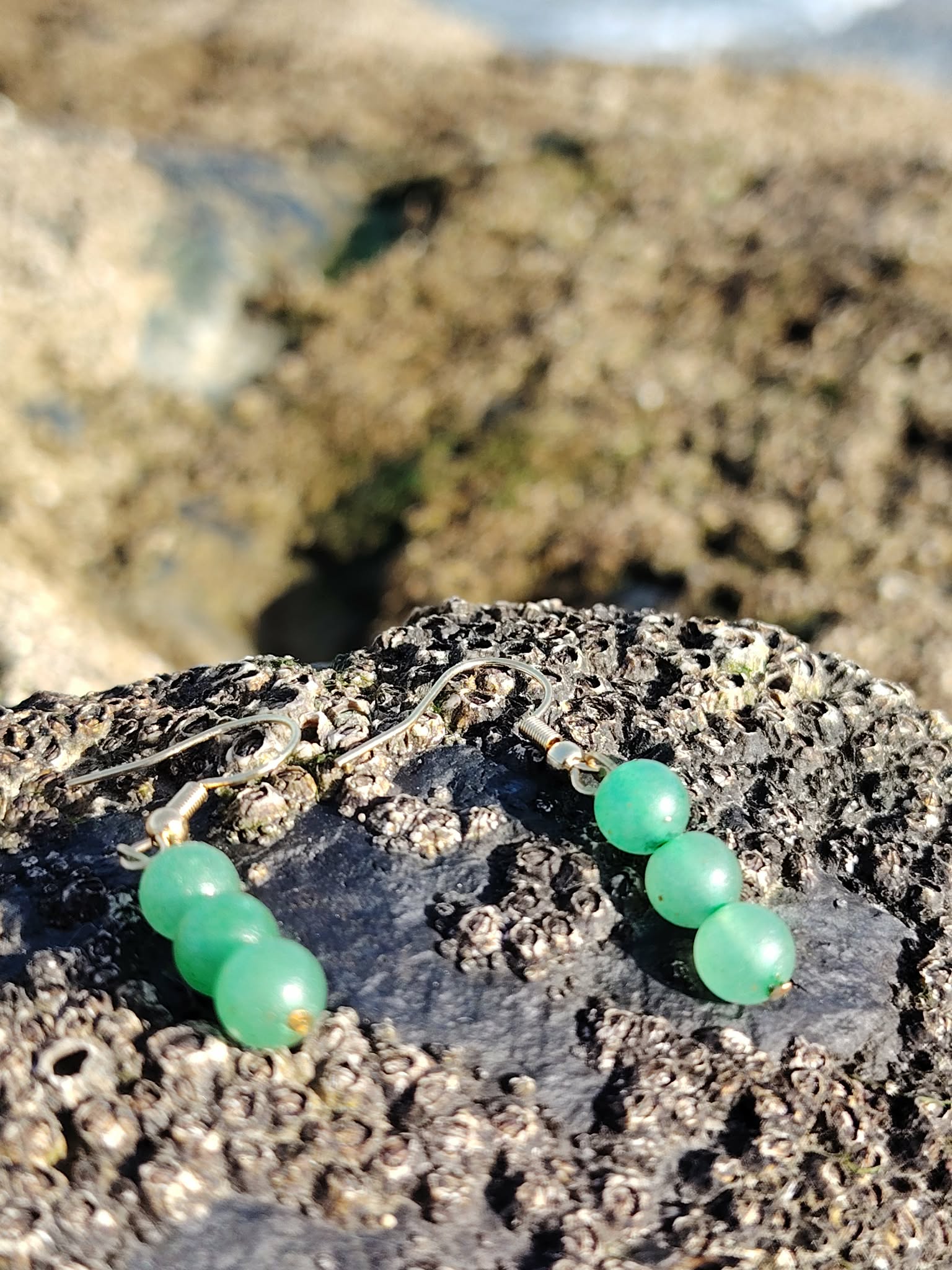 Boucles d’oreilles en aventurine verte, création Magic Gaïa, en gros plan sur rocher breton, ambiance bord de mer, pour apaisement et optimisme.