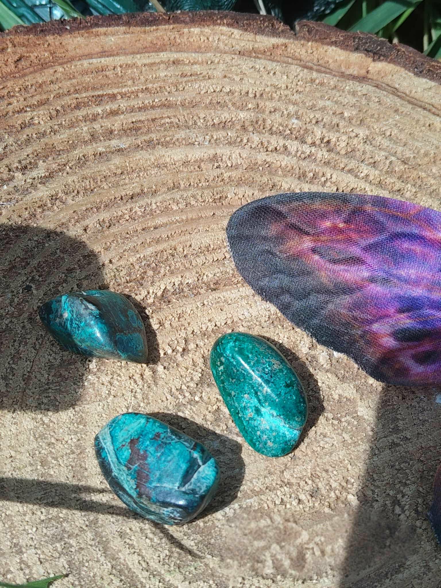 Trois galets de chrysocolle bleu-vert sur tranche de bois avec plume violette en décor, pierres roulées associées à la justesse intérieure, à l’inspiration paisible et à l’harmonie émotionnelle.