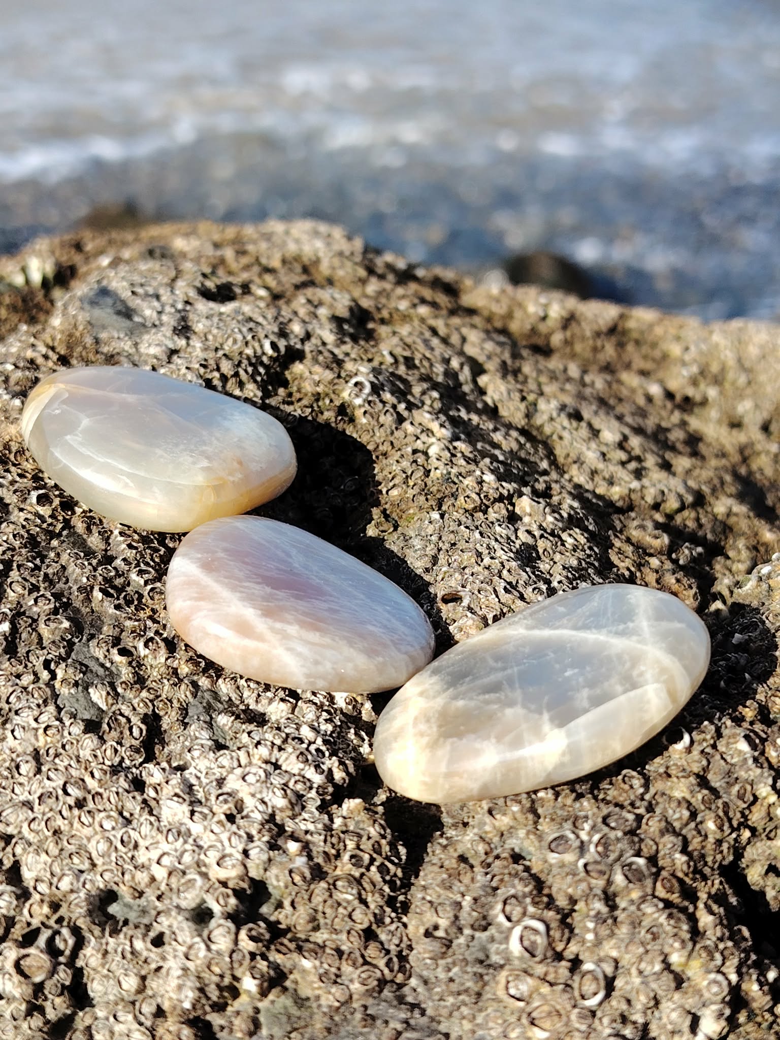 Trois galets de pierre de lune aux reflets nacrés posés sur un rocher face à la mer en Bretagne, pierres naturelles évoquant la fluidité intérieure, la rêverie et la tranquillité.