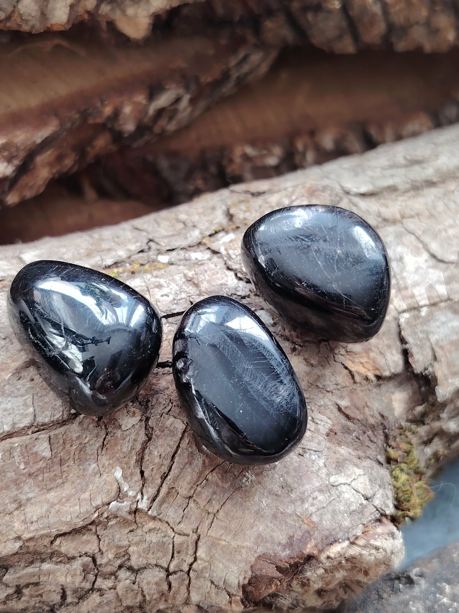 Trois pierres roulées d’hyperstène noir aux reflets métalliques posées sur du bois naturel, éclats subtils, pour inviter à l’ancrage, la protection et la clarté mentale.