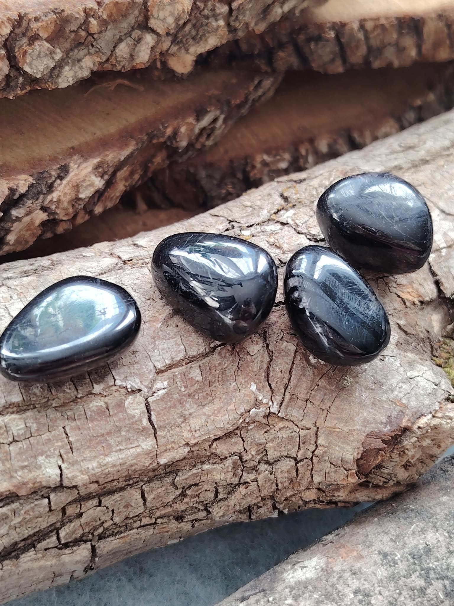 Quatre pierres roulées d’hyperstène noir aux reflets métalliques posées sur du bois craquelé, lumière douce, pour inviter à l’ancrage, la protection et la clarté mentale
