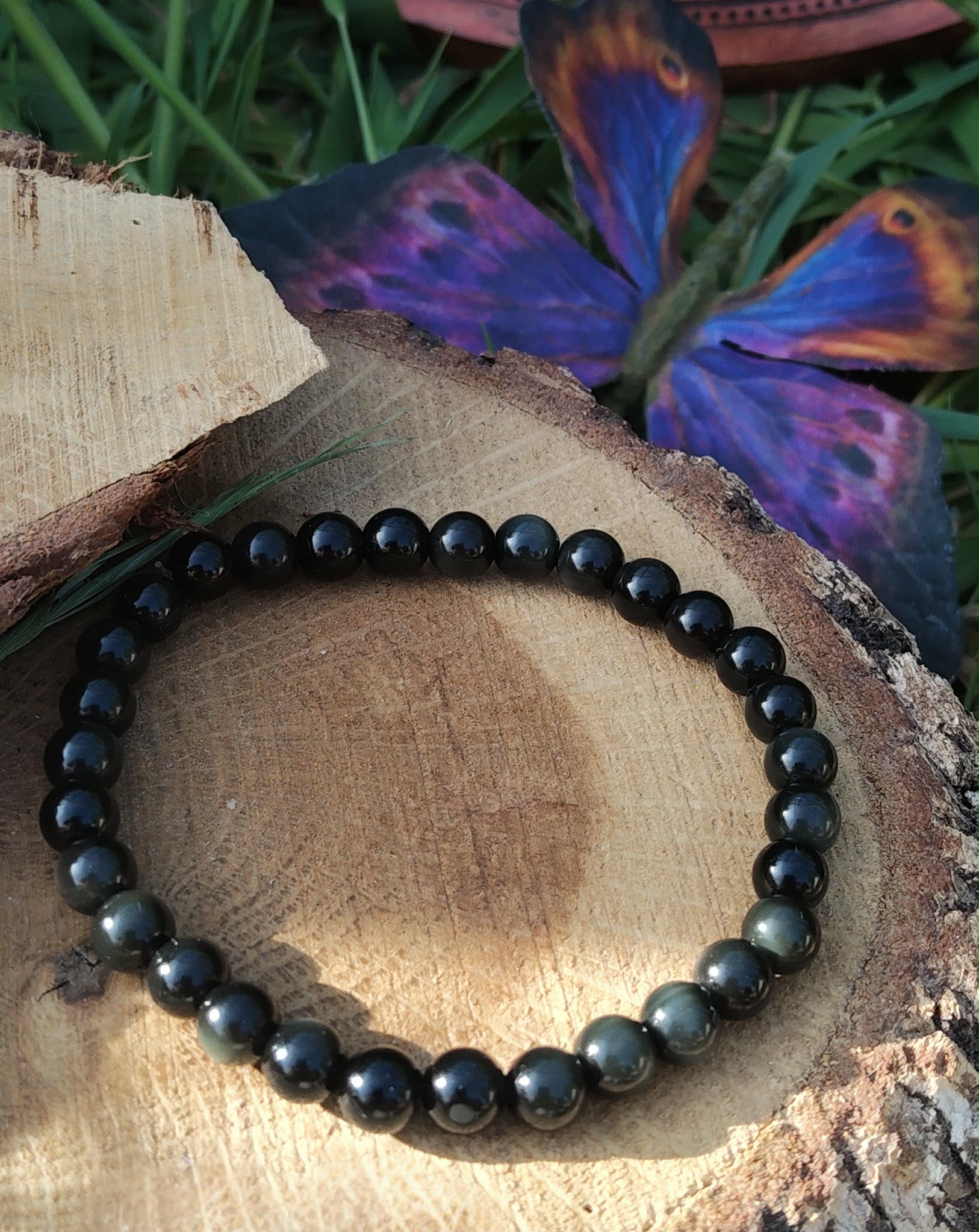 Bracelet obsidienne œil céleste 6 mm sur rondin de bois avec papillon bleu et brins d’herbe, perles noires irisées, introspection, recentrage et discernement."