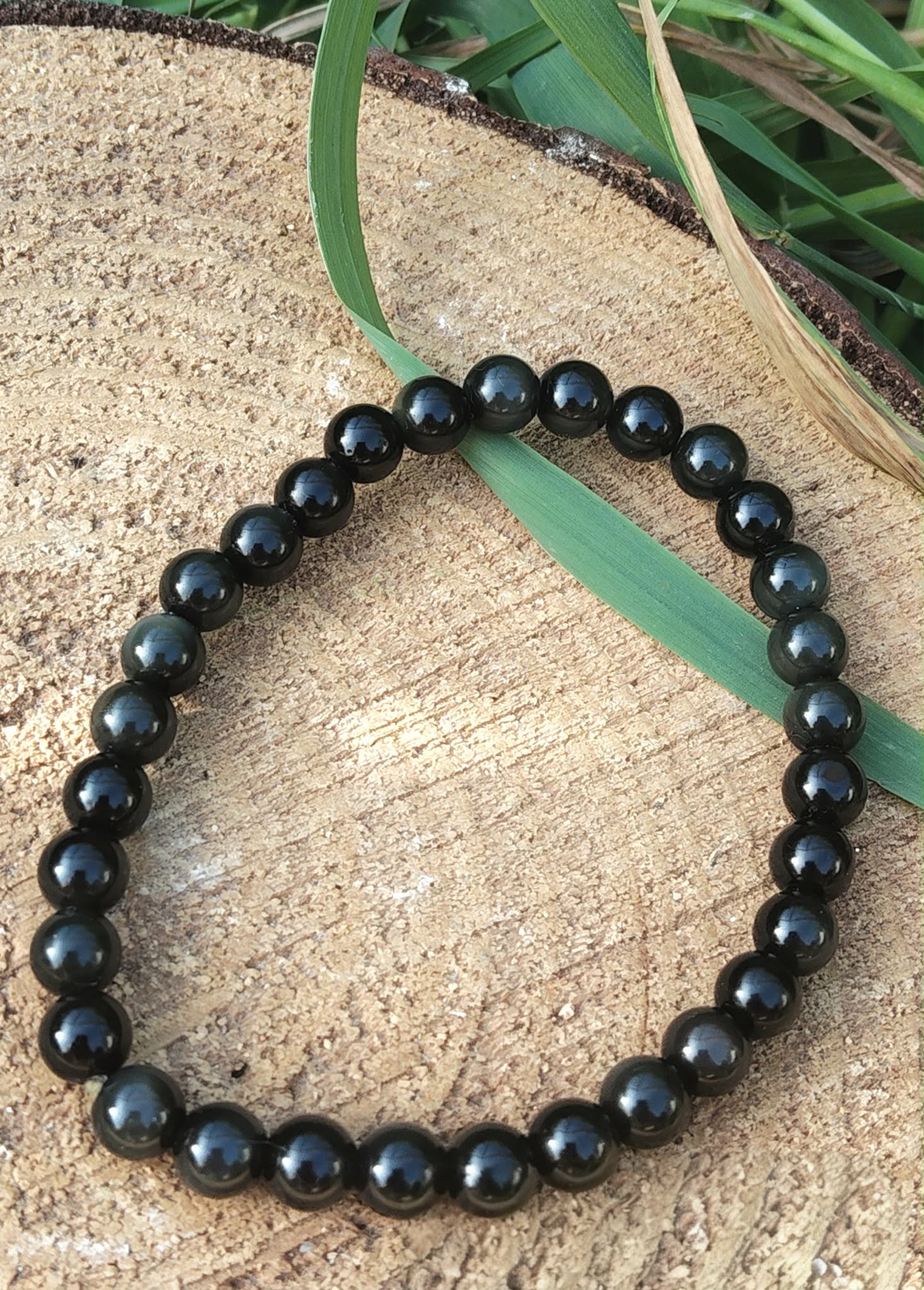 Bracelet obsidienne œil céleste 6 mm en perles noires sur rondin de bois avec brin d’herbe vert, assurance, présence et alignement personnel