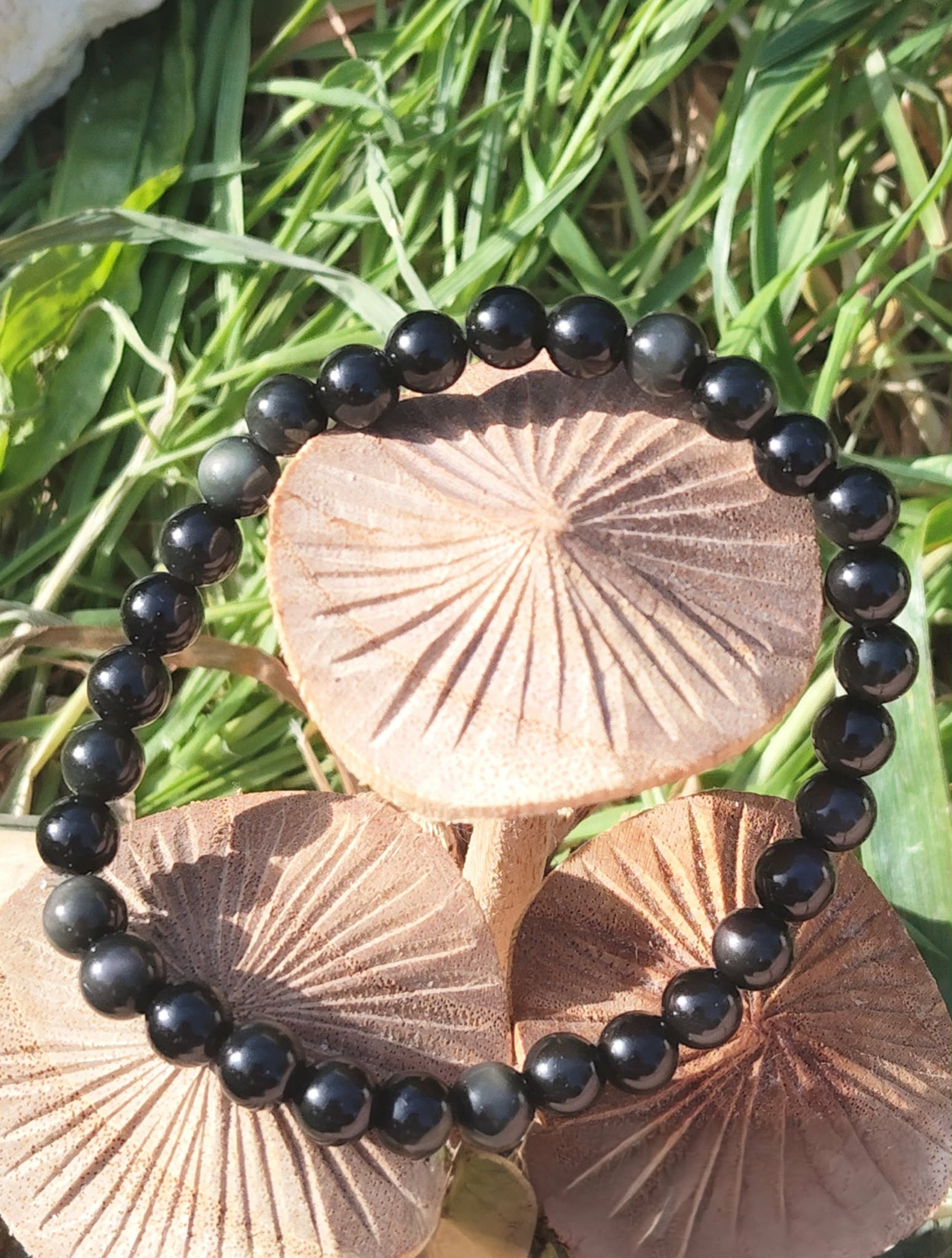 Bracelet obsidienne œil céleste 6 mm sur rondins en bois au milieu d’herbes vertes, perles noires, force calme, vigilance et enracinement