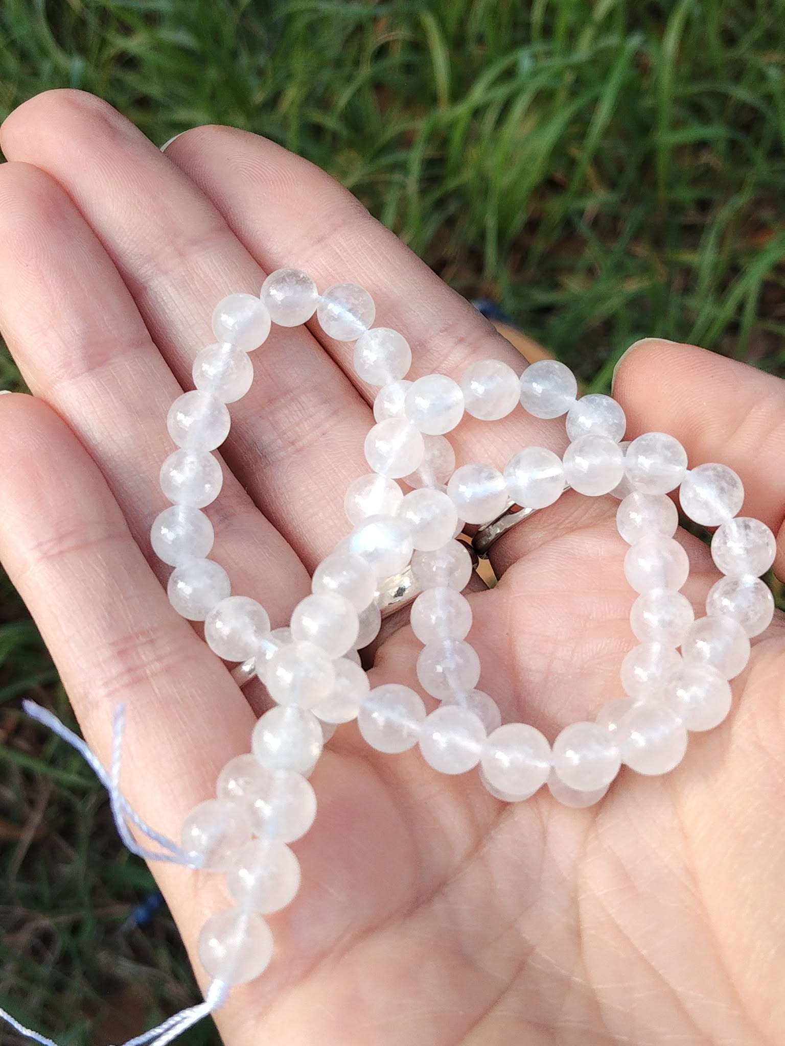 Fil de Perles de péristérite pierre de lune arc-en-ciel blanches aux reflets bleus dans la main sur herbe verte, bracelet 6 mm de calme et perception subtile.