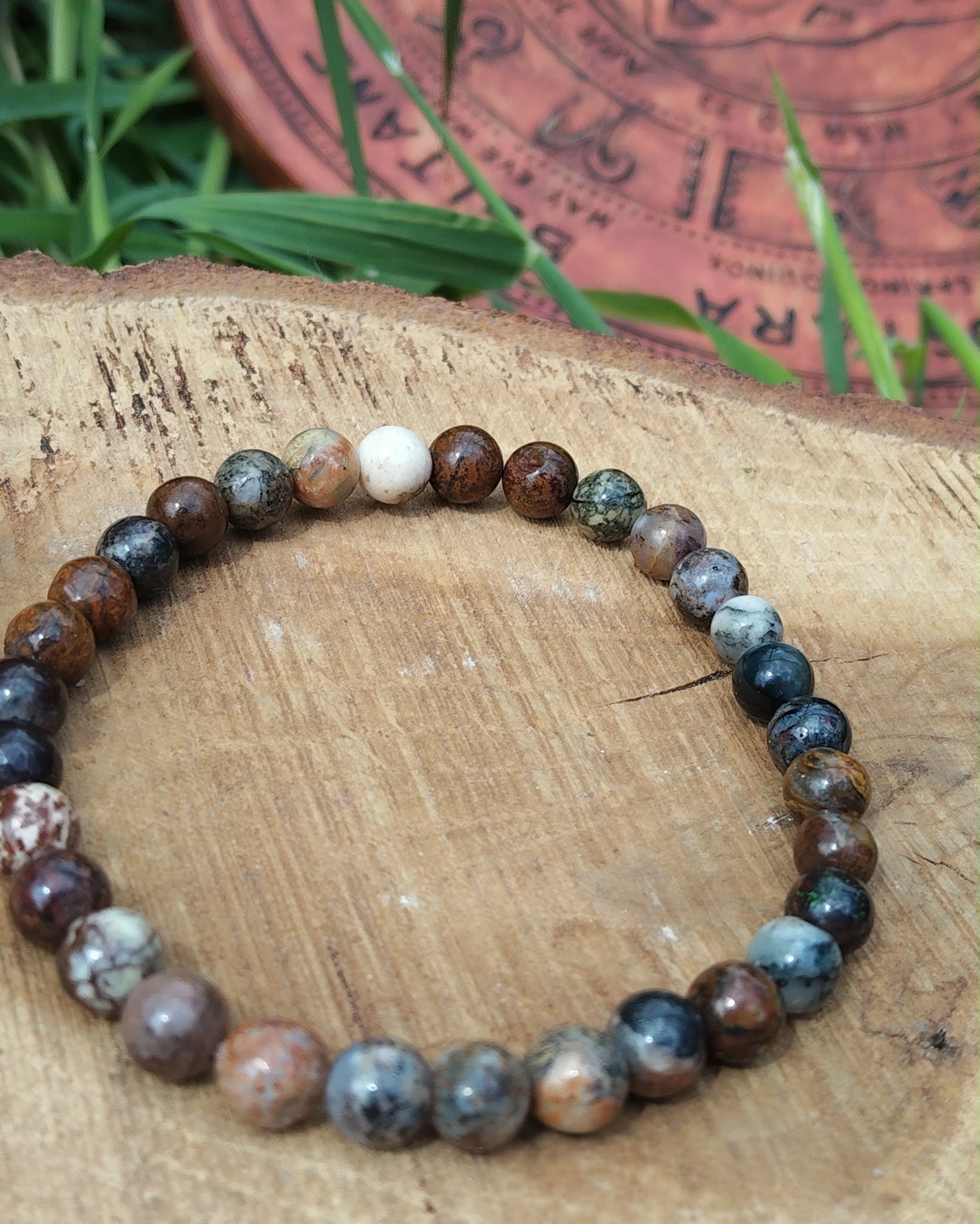 Bracelet Piétersite 6 mm création Magic Gaïa, perles brun-bleu nuancées en cercle sur rondin de bois gravé, herbes vertes et écorce en fond; apporte ancrage, clarté et courage.