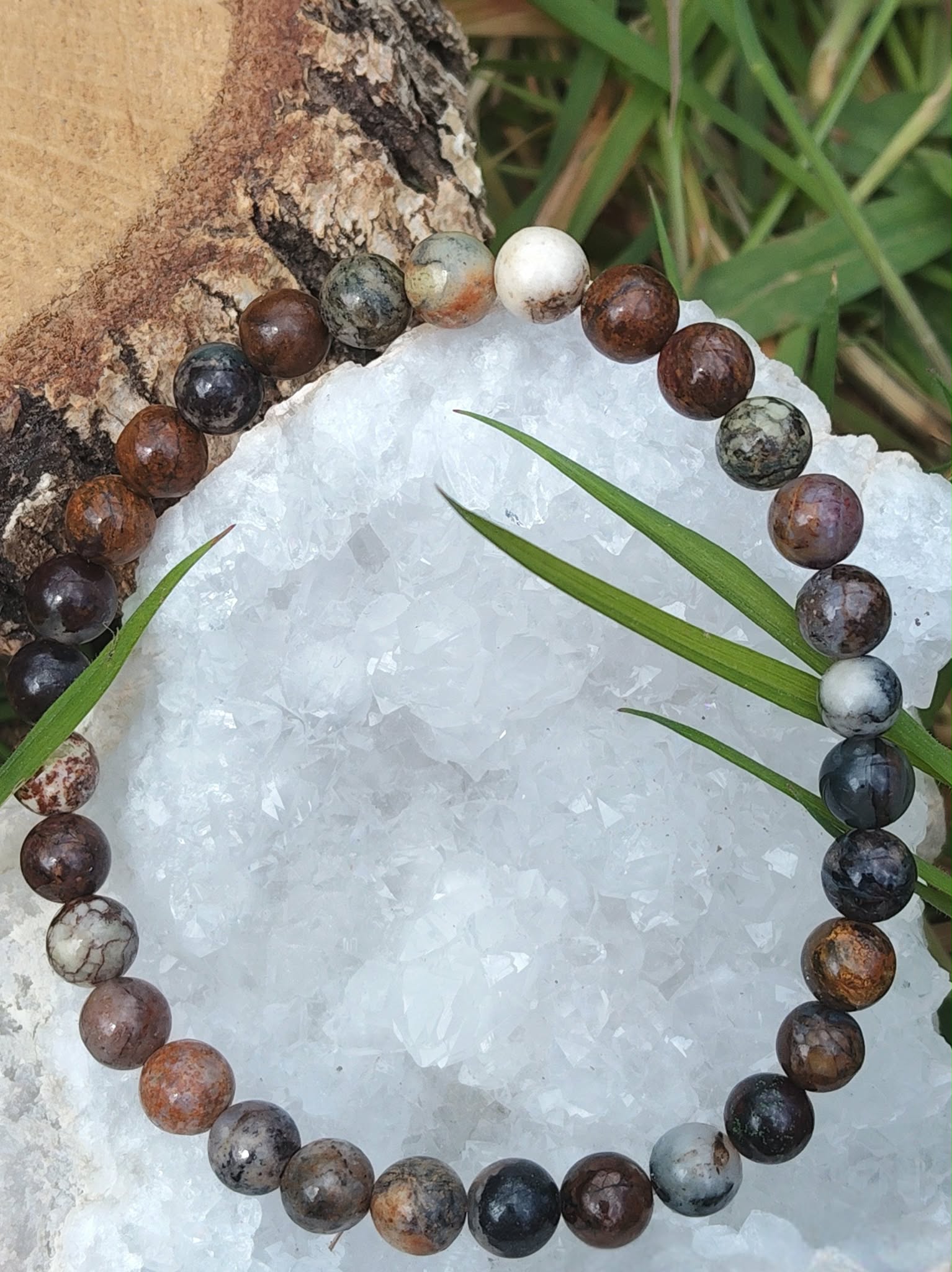 Bracelet en piétersite 6 mm aux perles bleu-brun posé sur une géode blanche, écorce et brins d’herbe en décor; ancrage, clarté mentale, confiance.