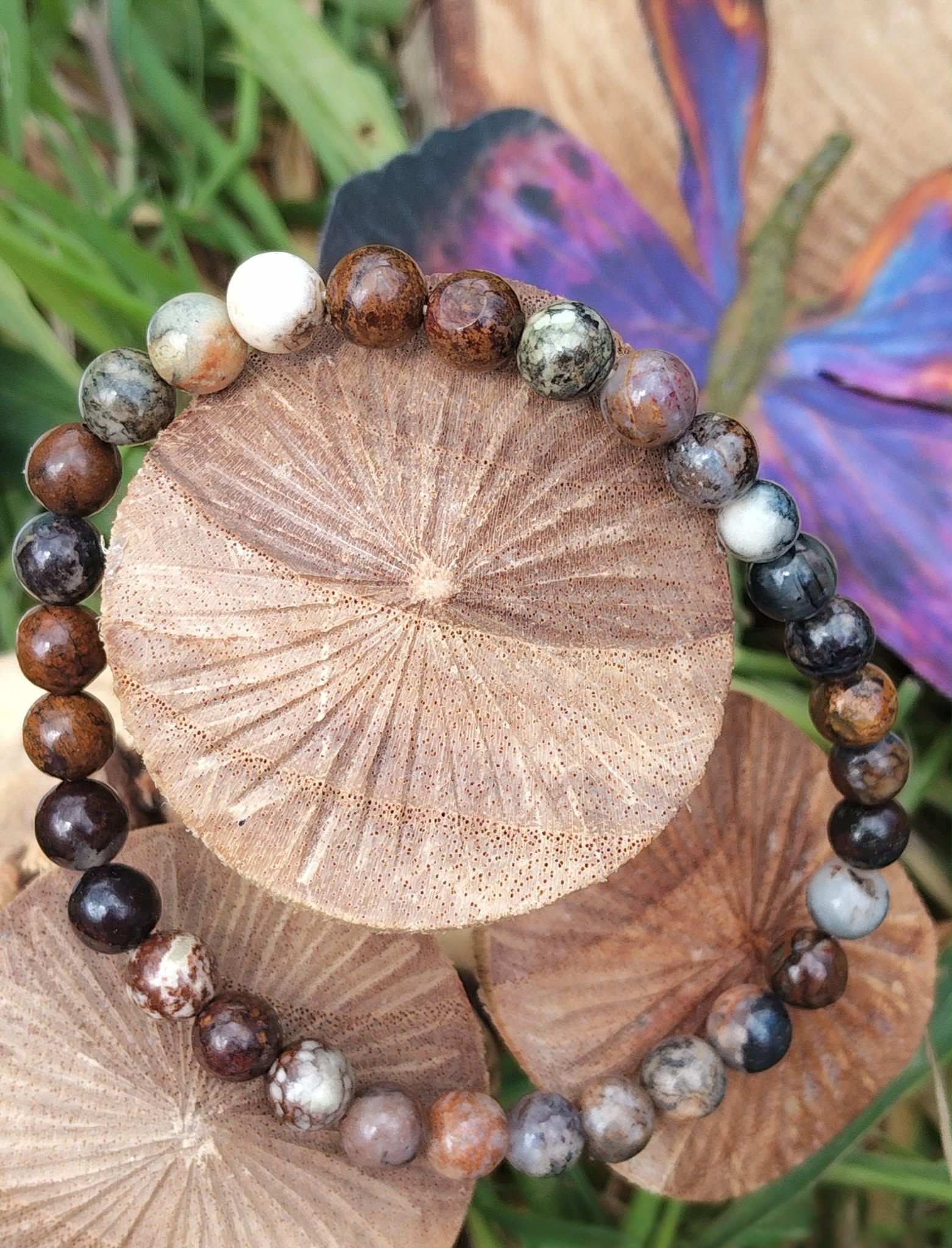Bracelet en piétersite 6 mm aux perles brun, bleu et or, posé sur rondin de bois gravé, herbes vertes et papillon violet en décor; apporte ancrage, clarté et confiance.