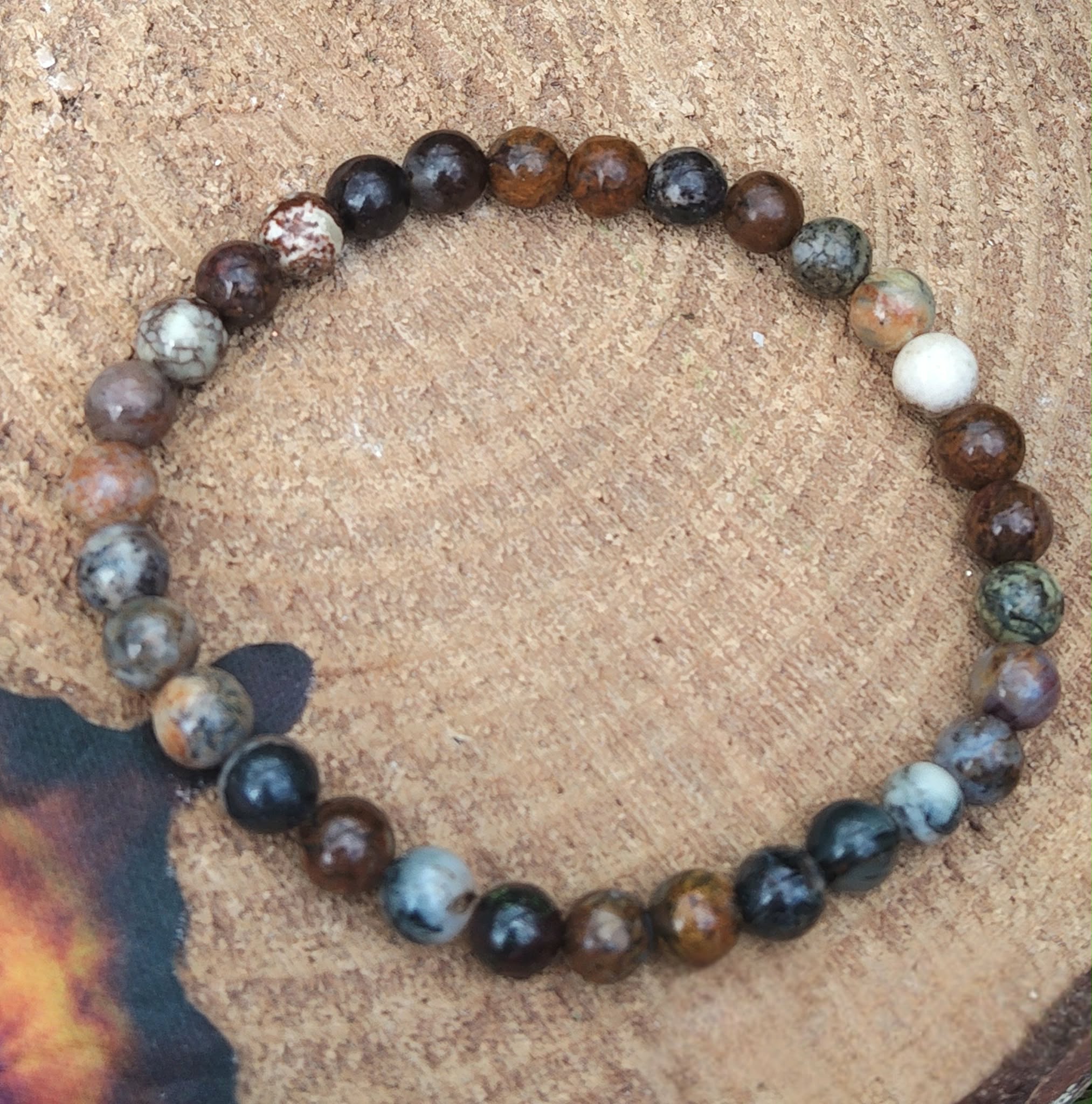 Bracelet piétersite 6 mm aux perles bleu pétrole, brun et doré, posé sur tissu sable près d’écorce et d’herbes vertes; invite à la clarté, au courage et à l’ancrage.