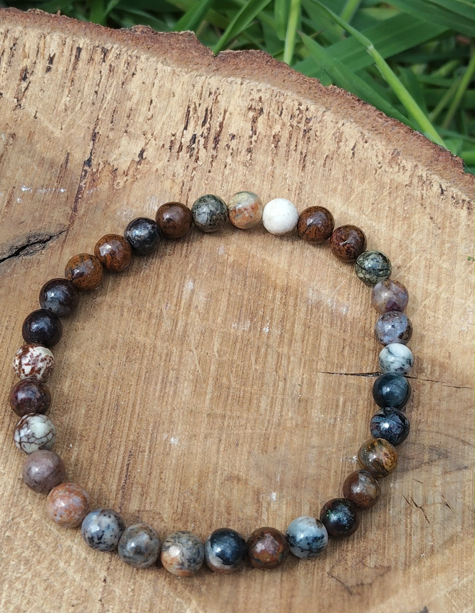 Bracelet Piétersite 6 mm en perles brun-bleu, posé en cercle sur rondin de bois gravé, brin d’herbe en arrière-plan; aide à l’ancrage, lucidité et confiance