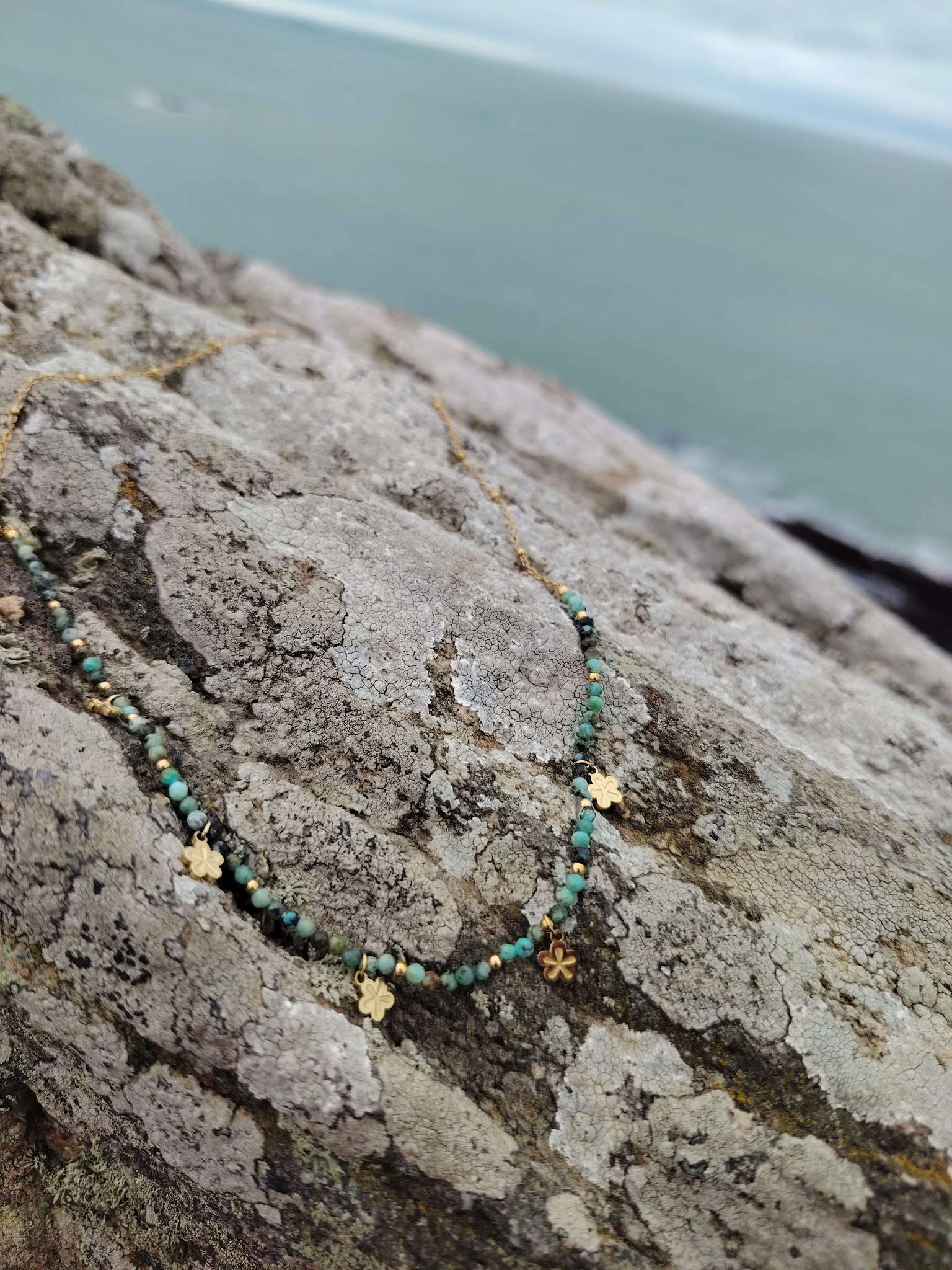 Collier doré à perles facettées vert africain posé sur rocher face à la mer en Bretagne, bijou fin au rendu frais et harmonieux