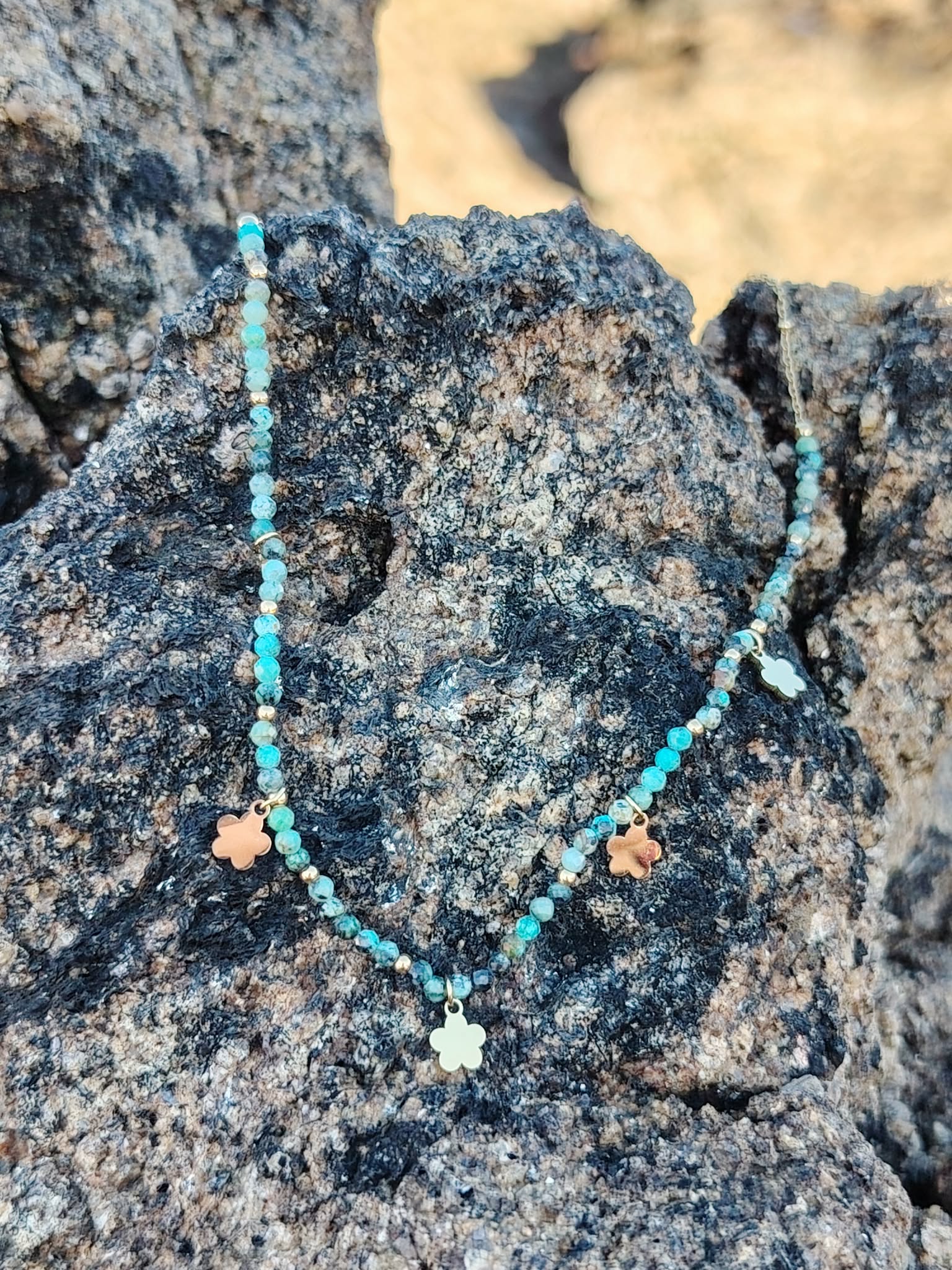 Collier acier inoxydable doré avec perles vert africain et pendentifs fleur photographié sur rochers bretons, élégance légère et esprit nature