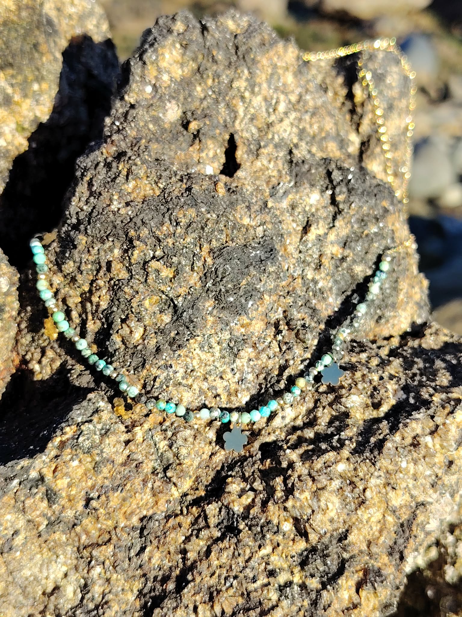 Collier doré en acier inoxydable avec perles vert africain facettées et pendentifs fleur, posé sur rocher marin pour une touche chic et printanière