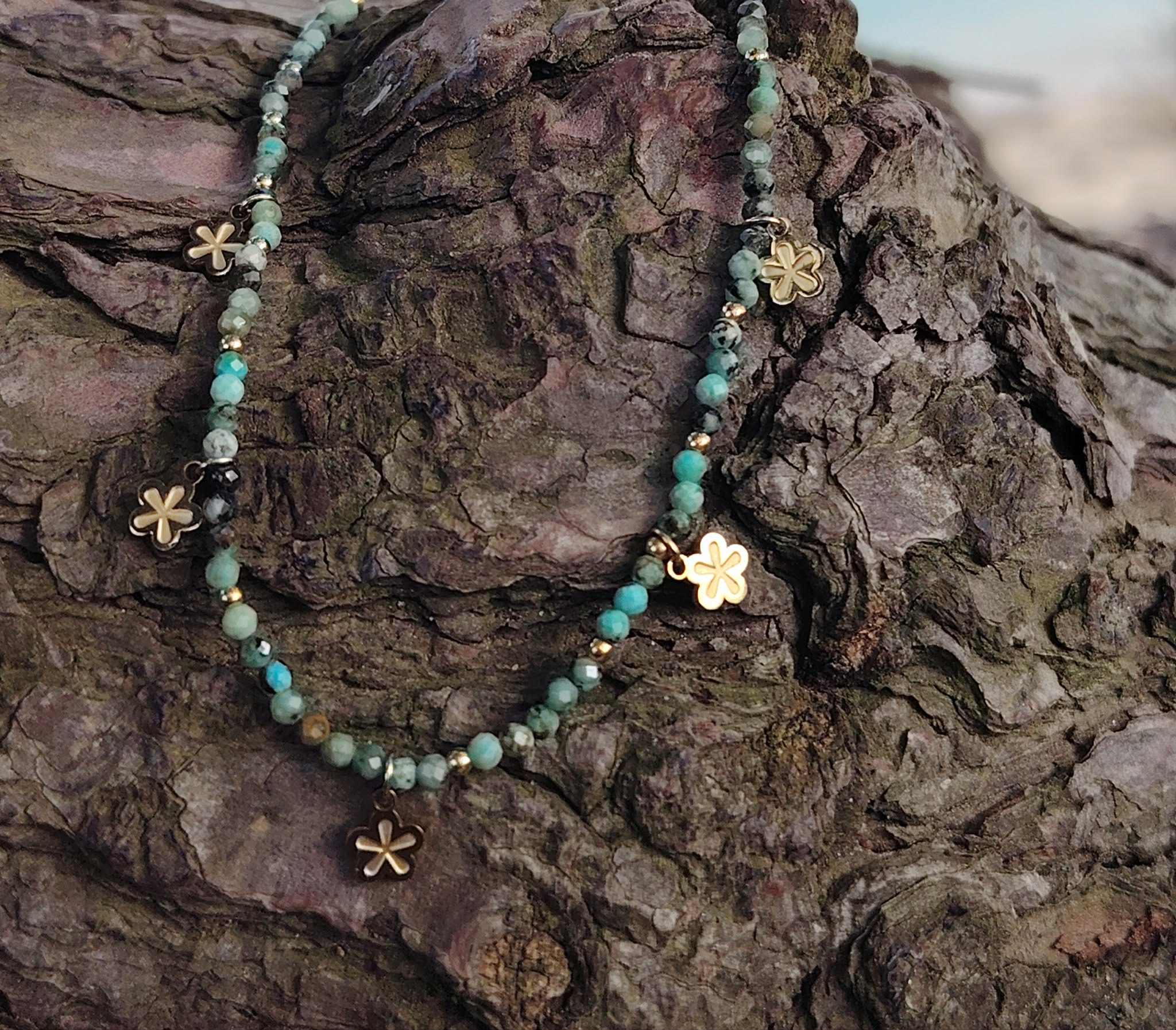 Gros plan sur collier doré en acier inoxydable avec perles facettées vert africain et pendentifs fleur, éclat lumineux sur rochers bruns