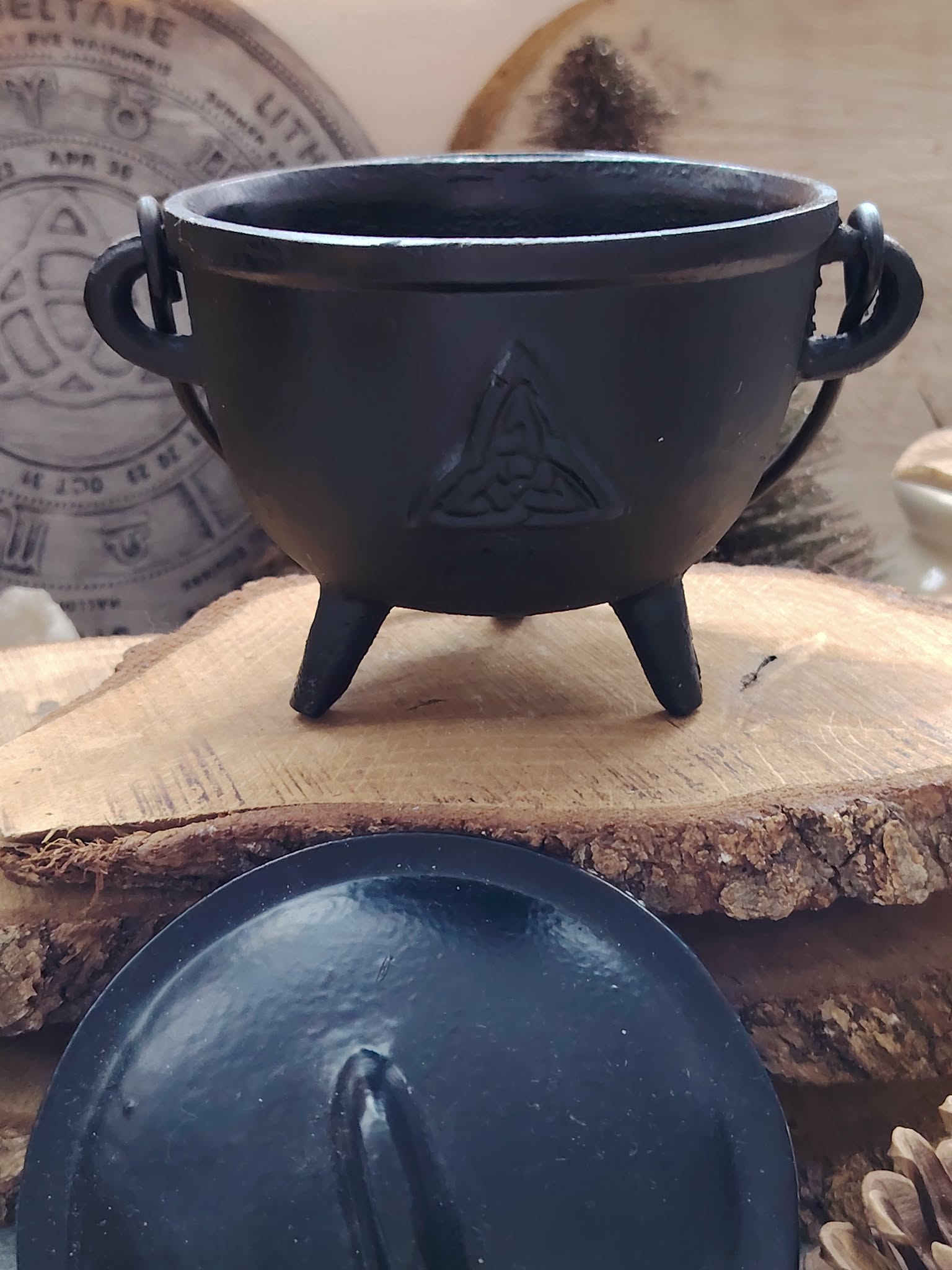 Petit chaudron en fonte noire avec trois pieds sur planche en bois, couvercle au premier plan, accessoire spirituel pour autel.