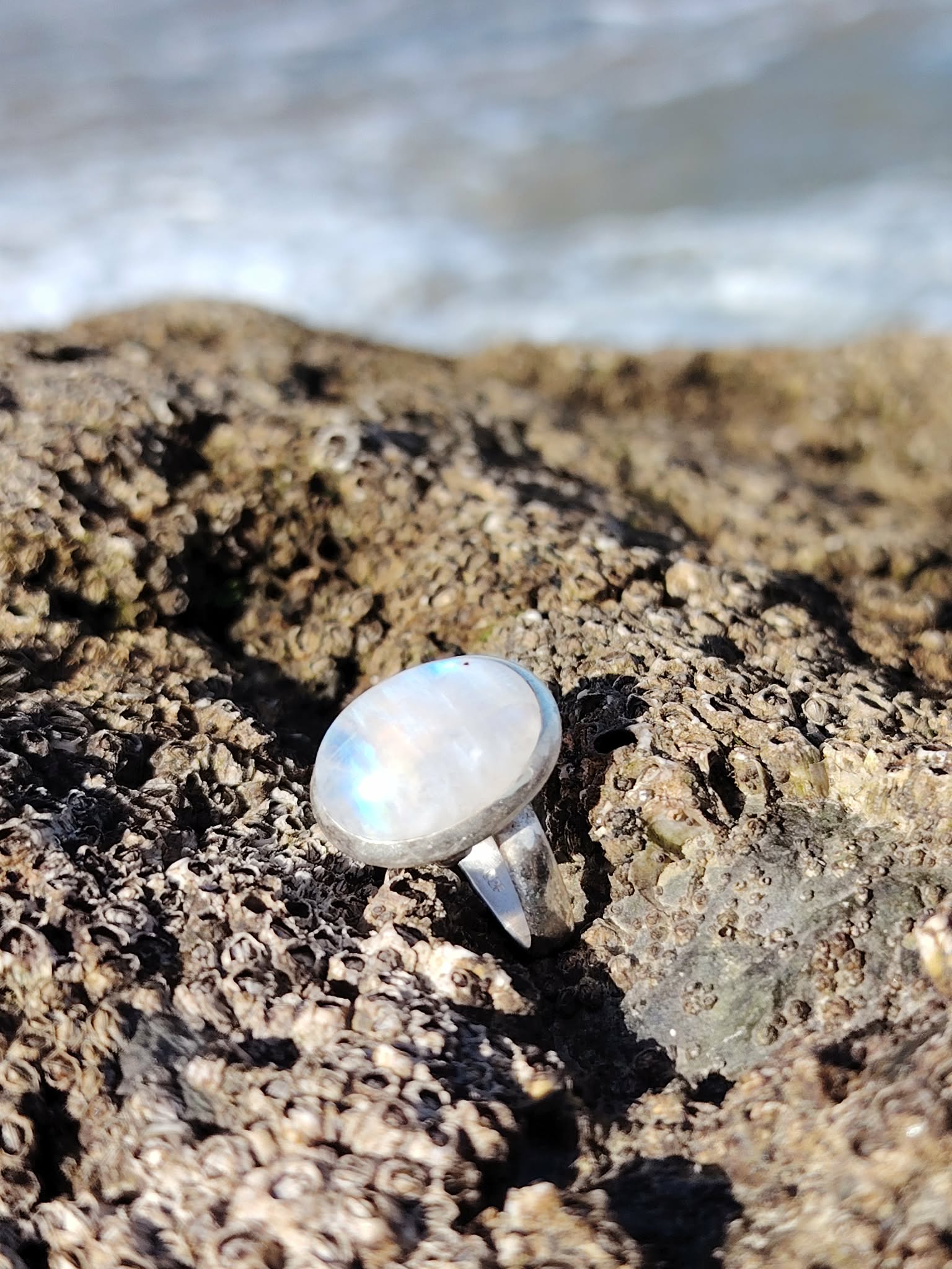 Bague réglable en argent 925 sertie d’une péristérite (pierre de lune arc-en-ciel) ovale, posée sur roche sombre en bord de mer, reflets bleutés, intention douceur et intuition.