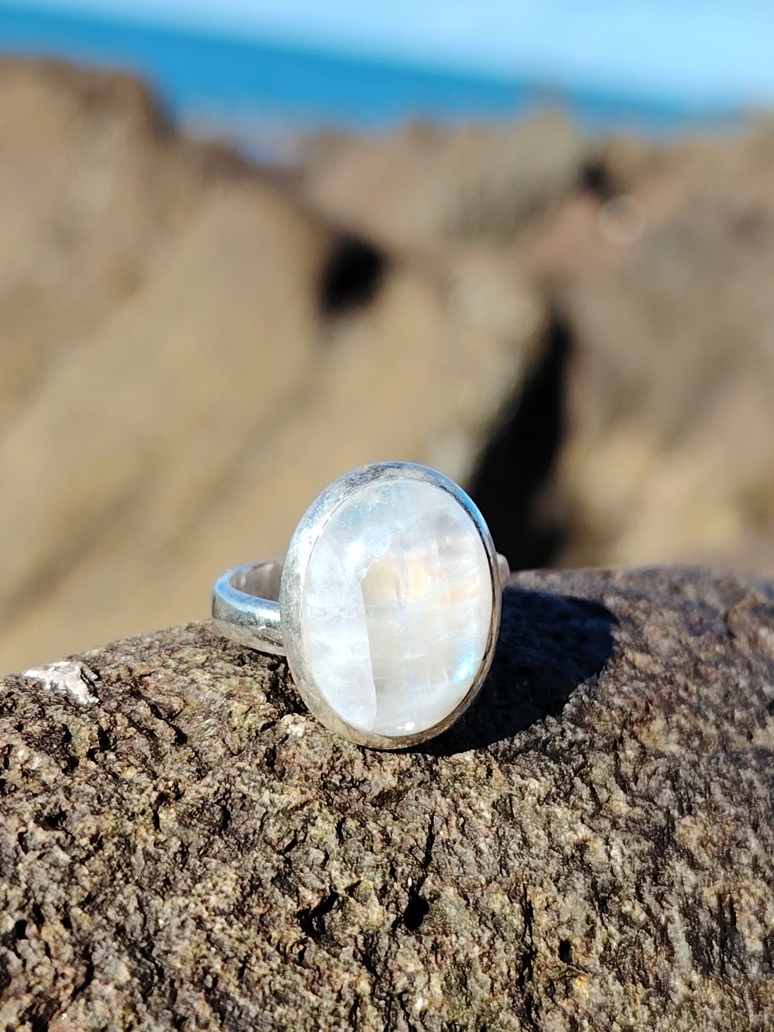 Bague réglable en argent 925 sertie d’une péristérite ovale (pierre de lune arc-en-ciel) posée sur roche granitée, mer de Bretagne floutée en arrière-plan, reflets laiteux, intention apaisement et intuition.