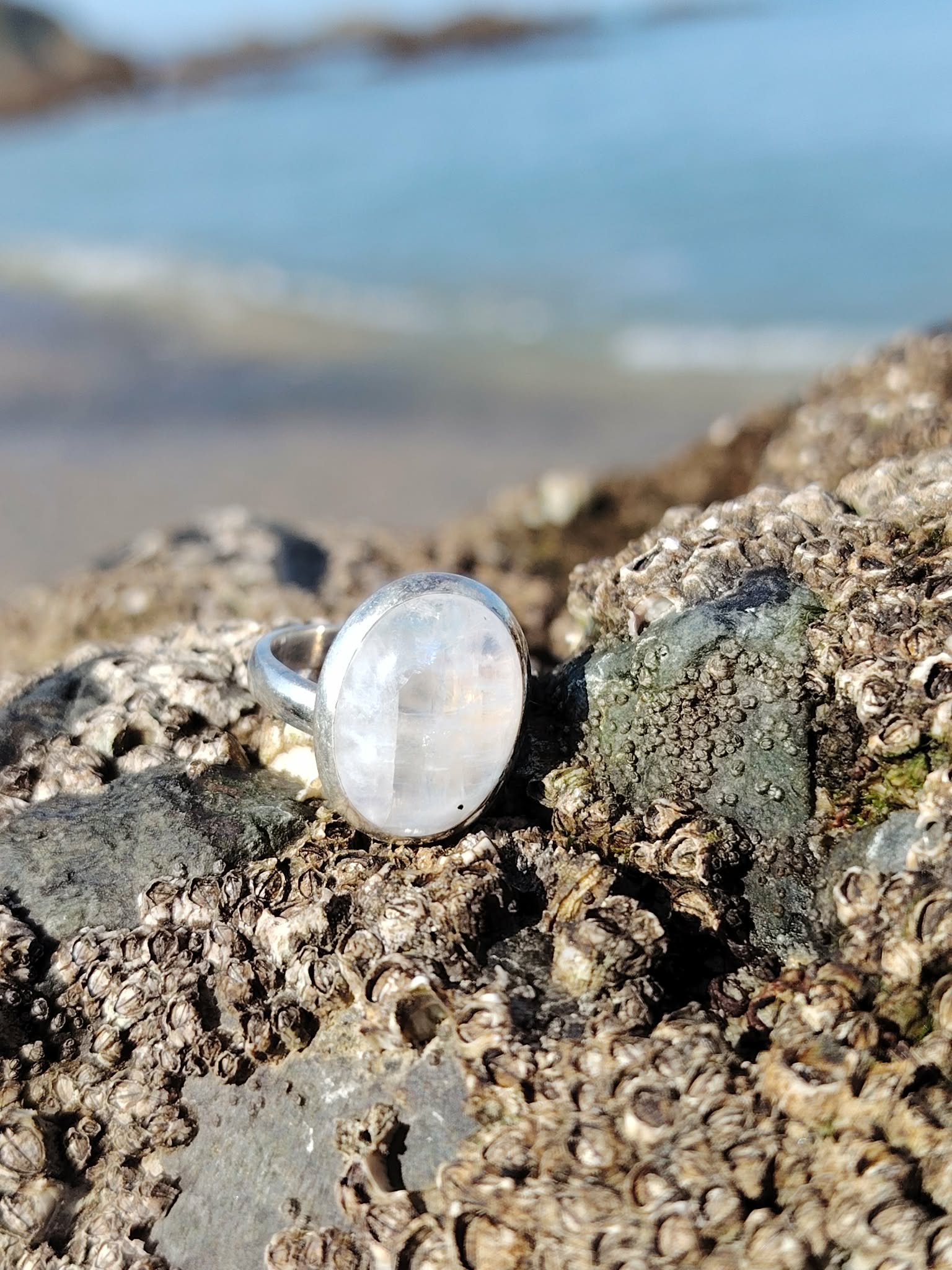 Bague réglable en argent 925 sertie d’une péristérite ovale (pierre de lune arc-en-ciel) posée sur rochers, plage et mer de Bretagne floutées en arrière-plan, reflets nacrés, intention apaisement et intuition
