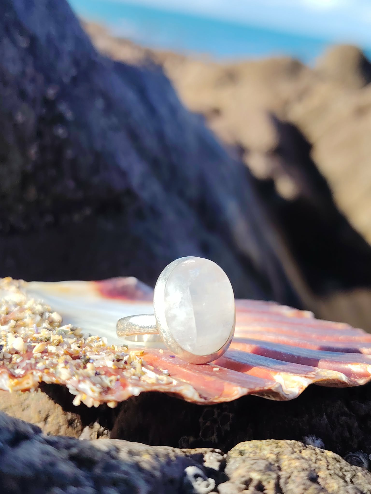 Bague réglable en argent 925 sertie d’une péristérite ovale (pierre de lune arc-en-ciel) posée sur coquillage et rochers en bord de mer, reflets laiteux, intention douceur et intuition.