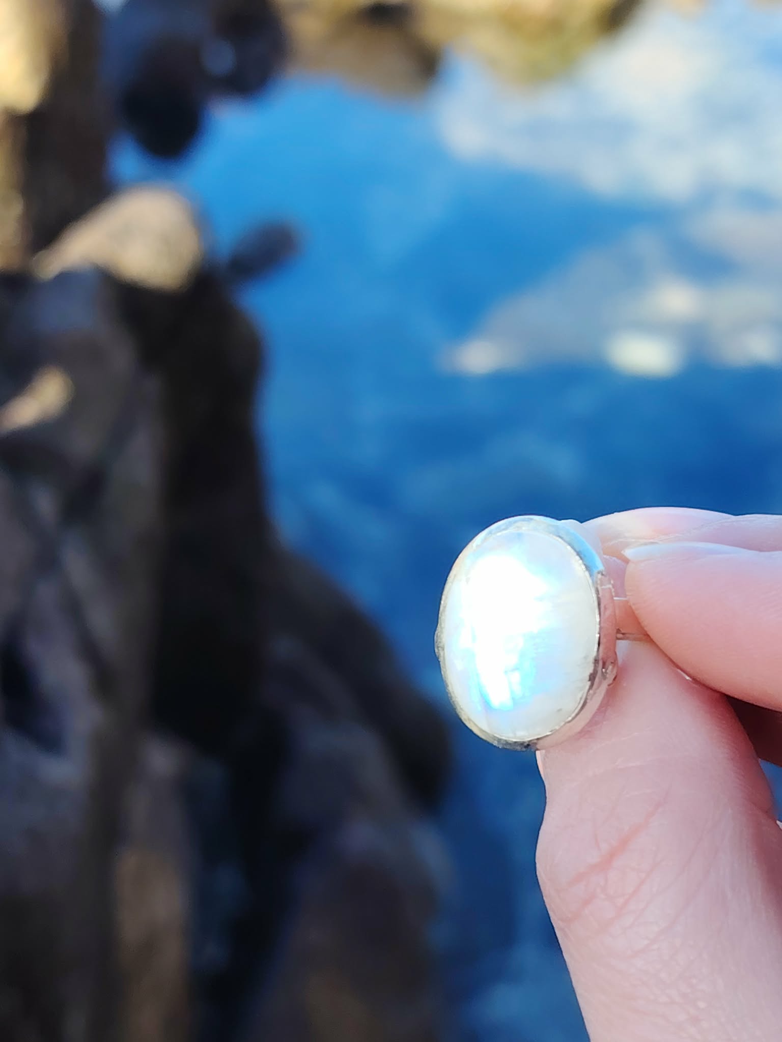 Bague réglable en argent 925 sertie d’une péristérite ovale (pierre de lune arc-en-ciel), tenue entre les doigts face à la mer de Bretagne, reflets bleutés, intention apaisement et intuition.