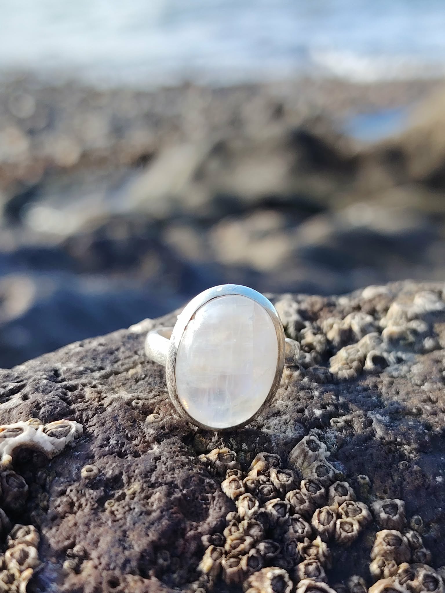 Bague réglable en argent 925 sertie d’une péristérite ovale (pierre de lune arc-en-ciel) posée sur roche granitée en bord de mer, reflets laiteux, intention apaisement et intuition.