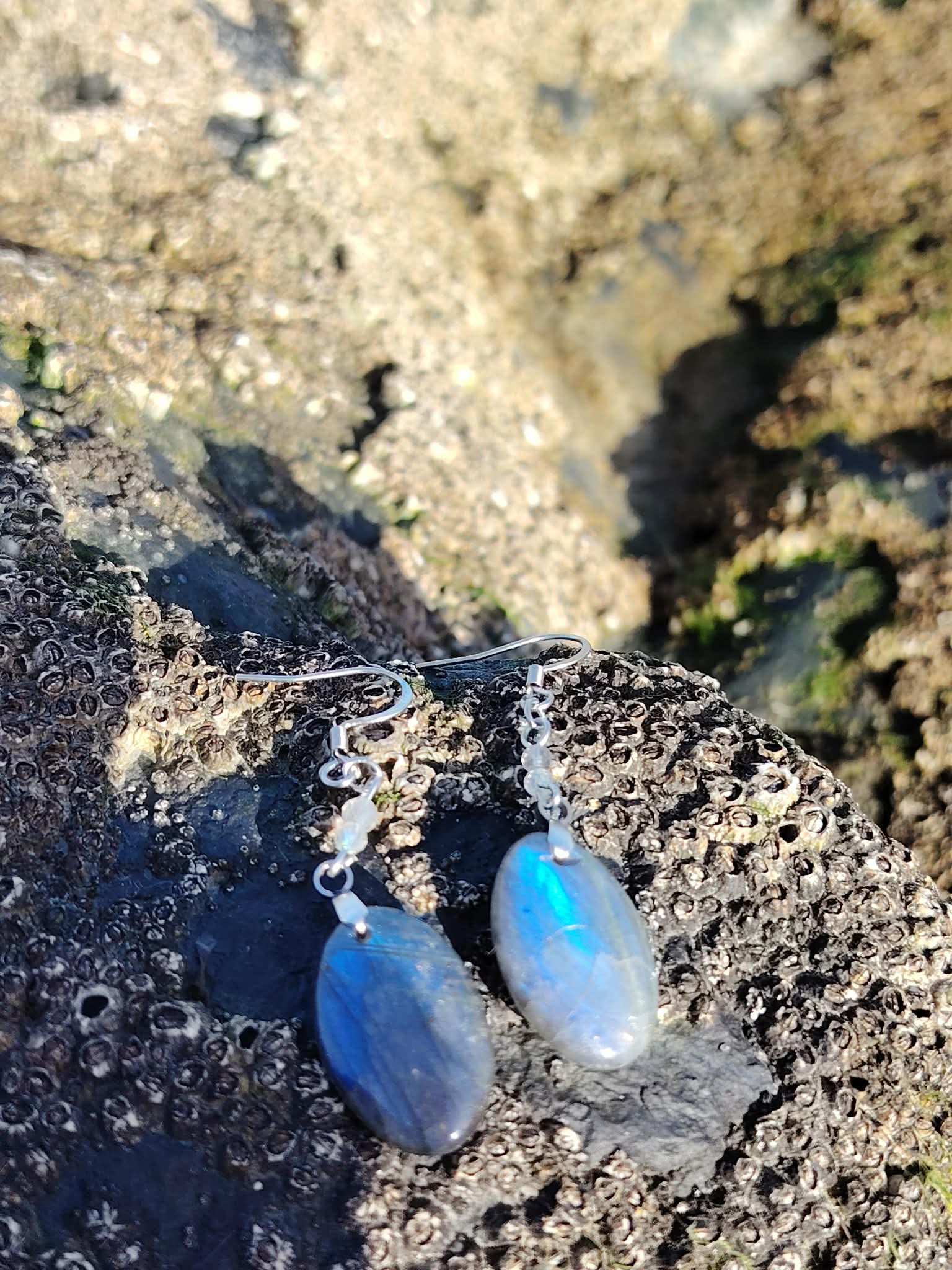 Boucles d’oreilles Labradorite création Magic Gaïa posées sur roche marine bretonne, reflets bleus au soleil; intention: protection douce, intuition, apaisement.