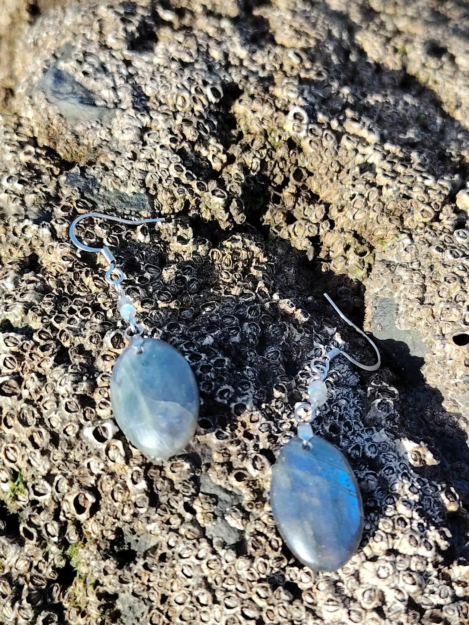 Boucles d’oreilles Labradorite création Magic Gaïa posées sur galets humides au bord de mer bretonne; intention: bouclier émotionnel, clarté, calme.