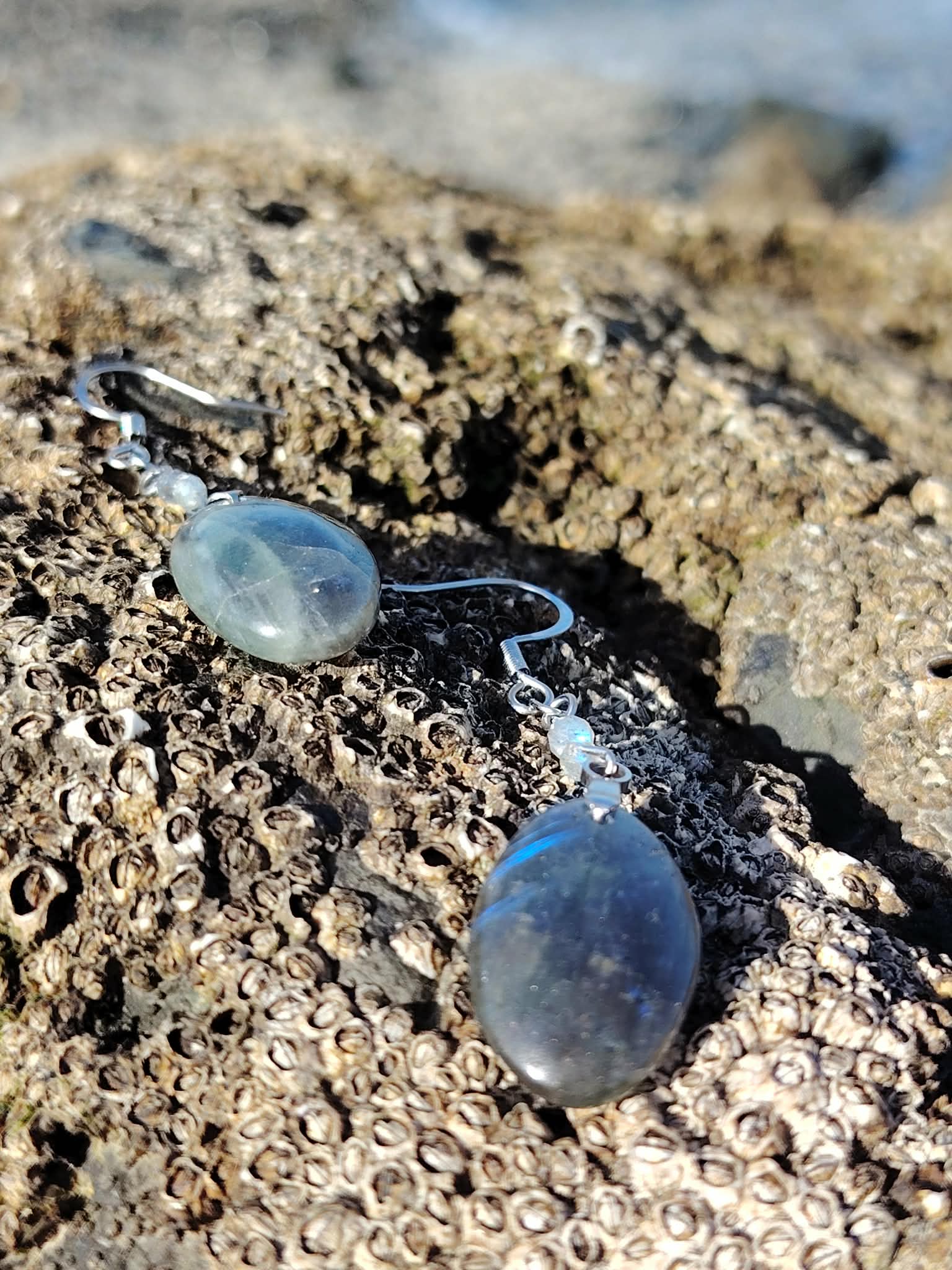 Boucles d’oreilles Labradorite création Magic Gaïa posées sur rocher breton couvert de bernacles, mer bleue en arrière-plan, reflets protecteurs et apaisants.
