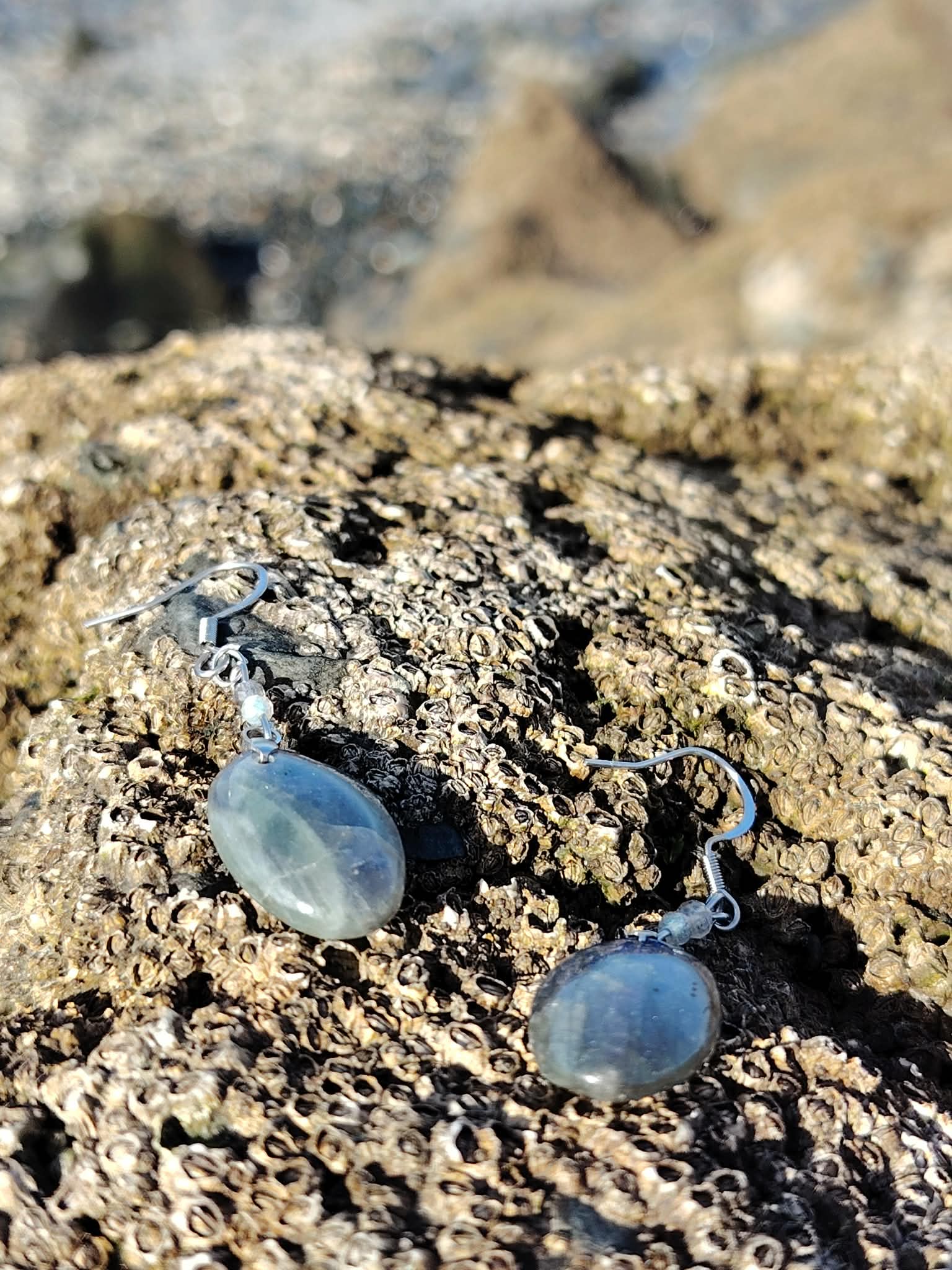 Boucles d’oreilles Labradorite création Magic Gaïa sur algues et sable doré, écume floue en fond; intention: ancrage léger, sérénité, confiance.