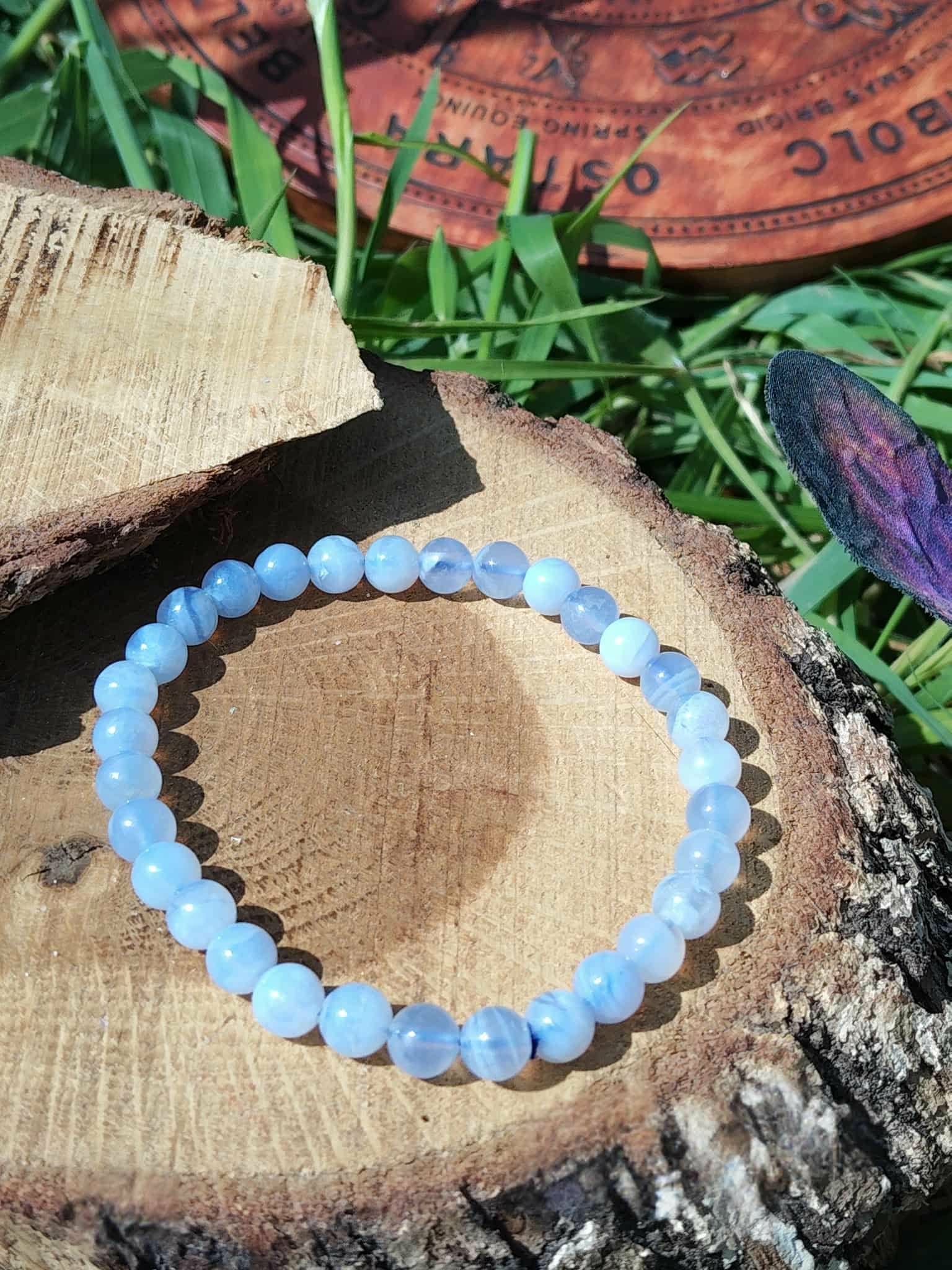 Bracelet calcédoine bleue 6 mm sur coupe de bois brut au soleil, herbe haute, disque gravé et plume violette, douceur relationnelle et expression sereine.