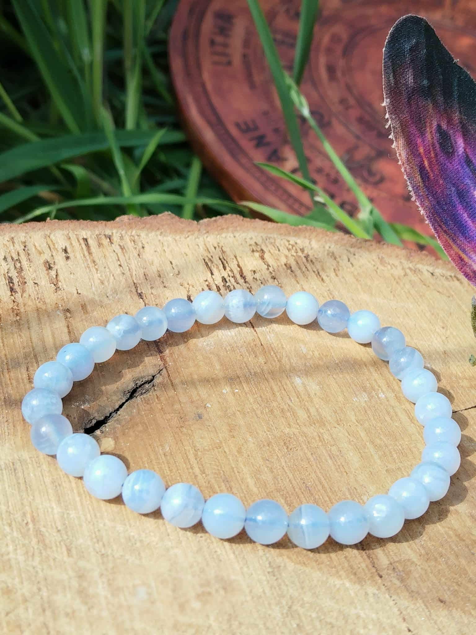 Bracelet en calcédoine bleue 6 mm sur rondin de bois clair au soleil, herbe verte, disque gravé et plume irisée, sérénité intérieure et clarté dans l’échange.