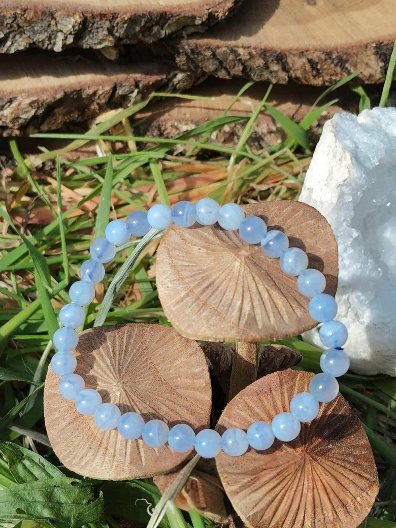 Bracelet calcédoine bleue 6 mm posé sur champignons en bois dans l’herbe avec écorce et géode de quartz, écoute paisible et stabilité affective